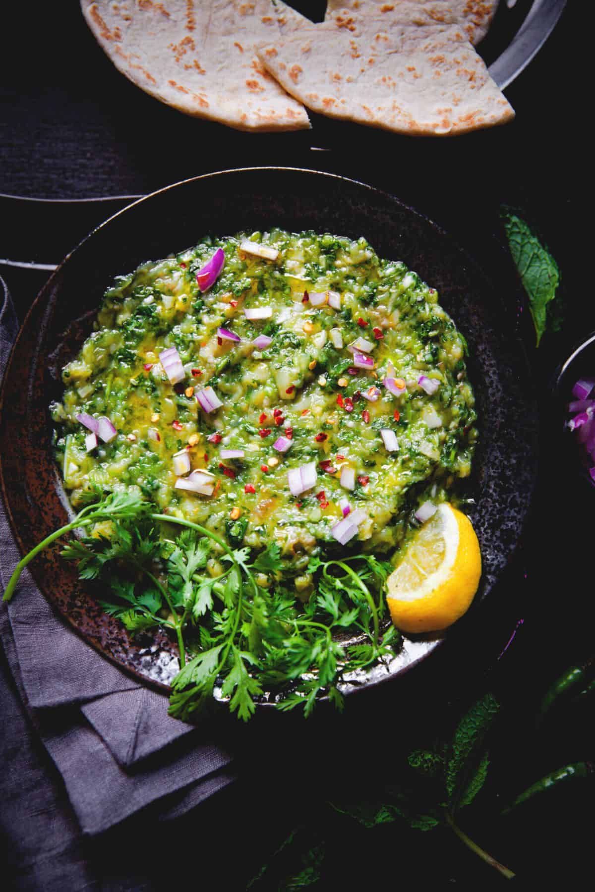 Green baingan bharta garnished with lemon wedge and cilantro served in a black plate.