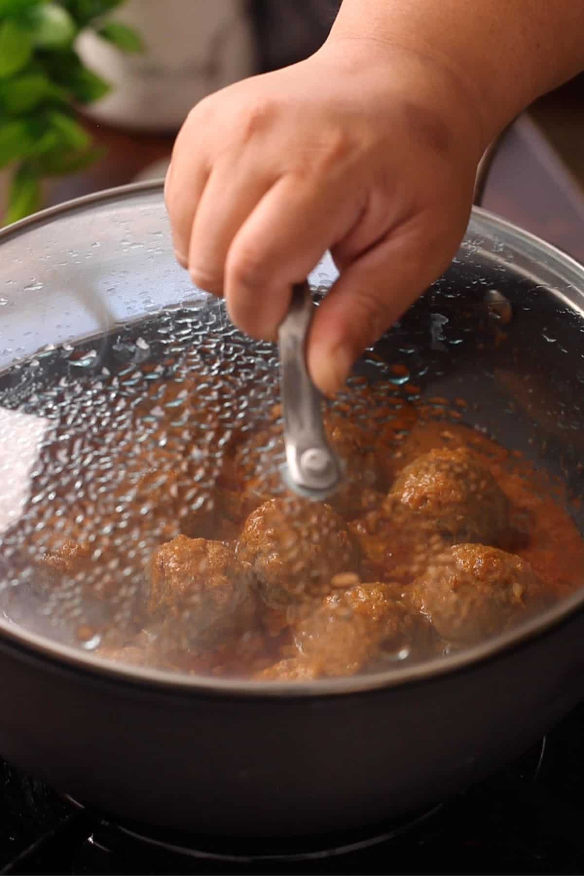 A hand puts the lid on a pan filled with simmering kofta curry in on a stovetop, with condensation on the glass lid.