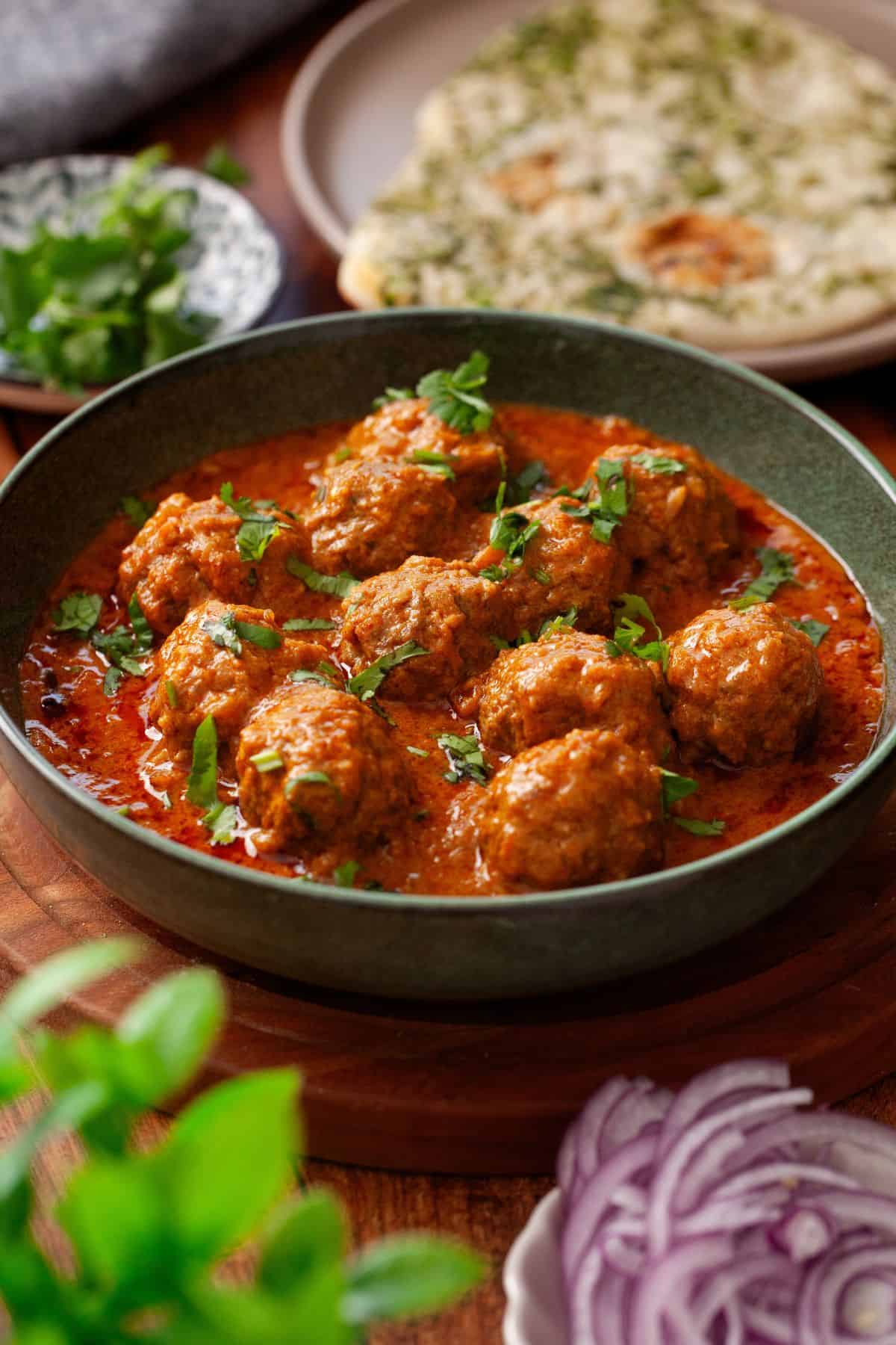 A bowl of kofta curry in a garnished with fresh cilantro. In the background, there is naan, herbs, and a plate of sliced red onions on a wooden surface.