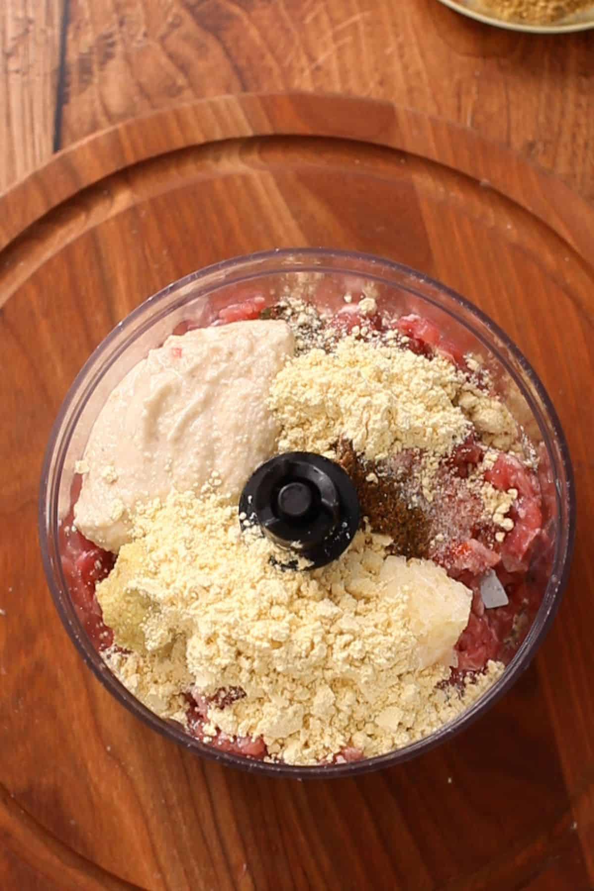 A food processor bowl filled with raw ground meat, gram flour, cashew paste, and various spices, all on a round wooden cutting board.