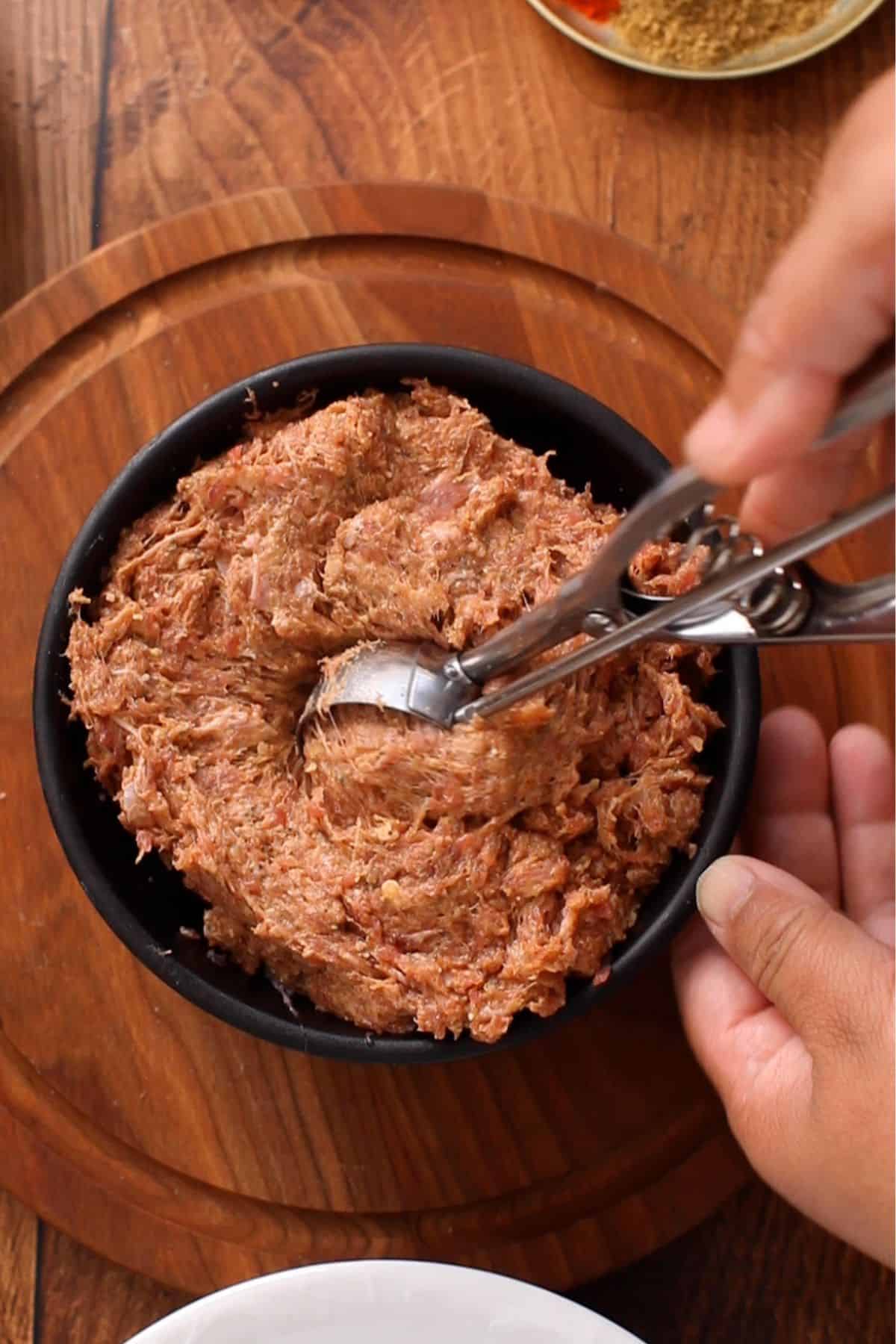 Using a scoop to portion kofta meat mixture from a black bowl placed on a wooden cutting board.