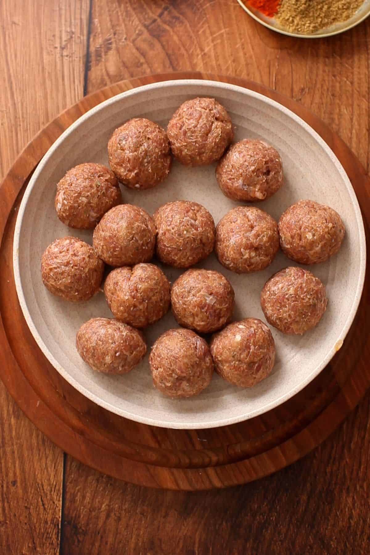 A round plate on a wooden surface holding fifteen raw kofta meatballs, arranged in a single layer. 