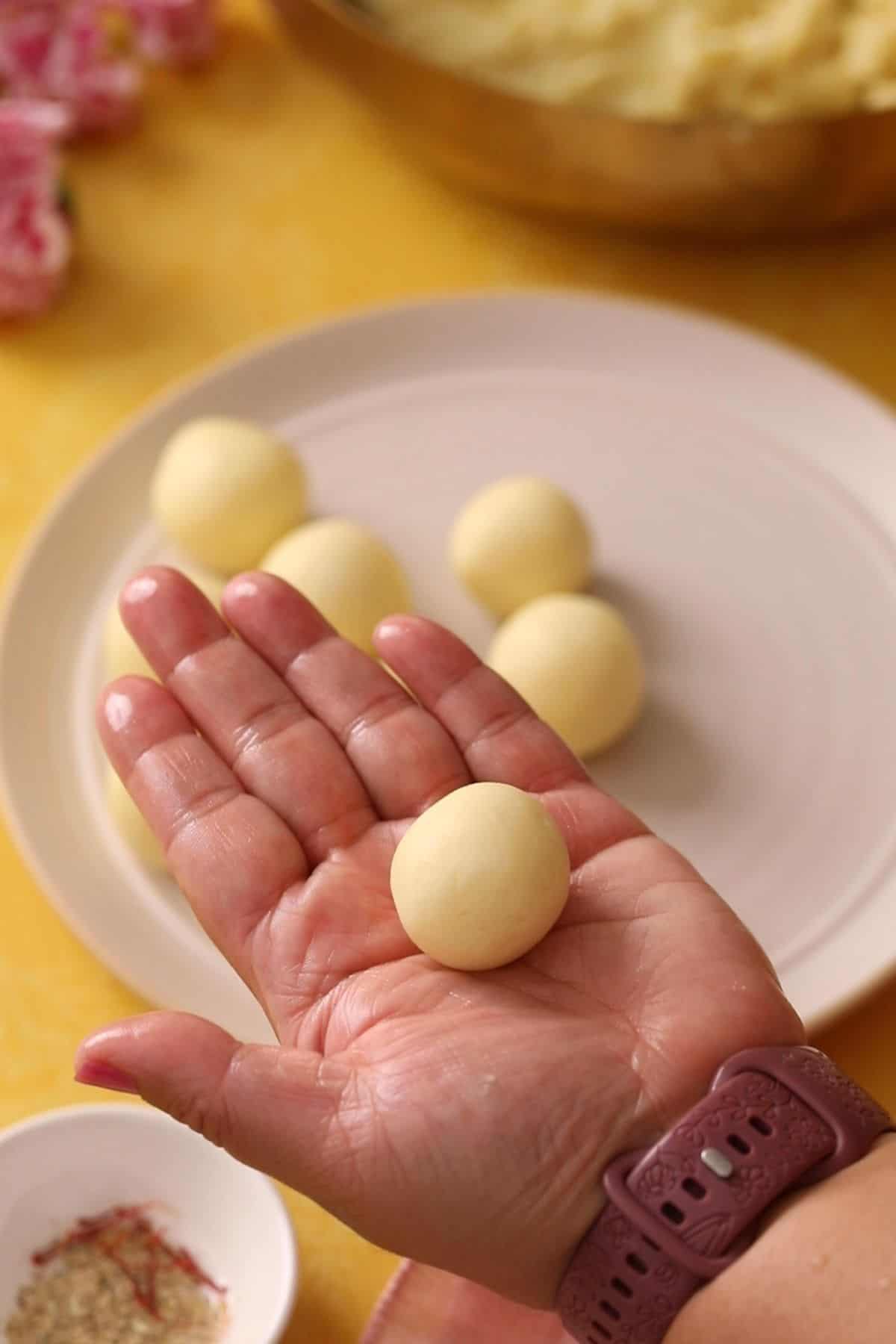 A hand holds a small round ball of gulab jamun dough, with several similar dough balls on a plate in the background.