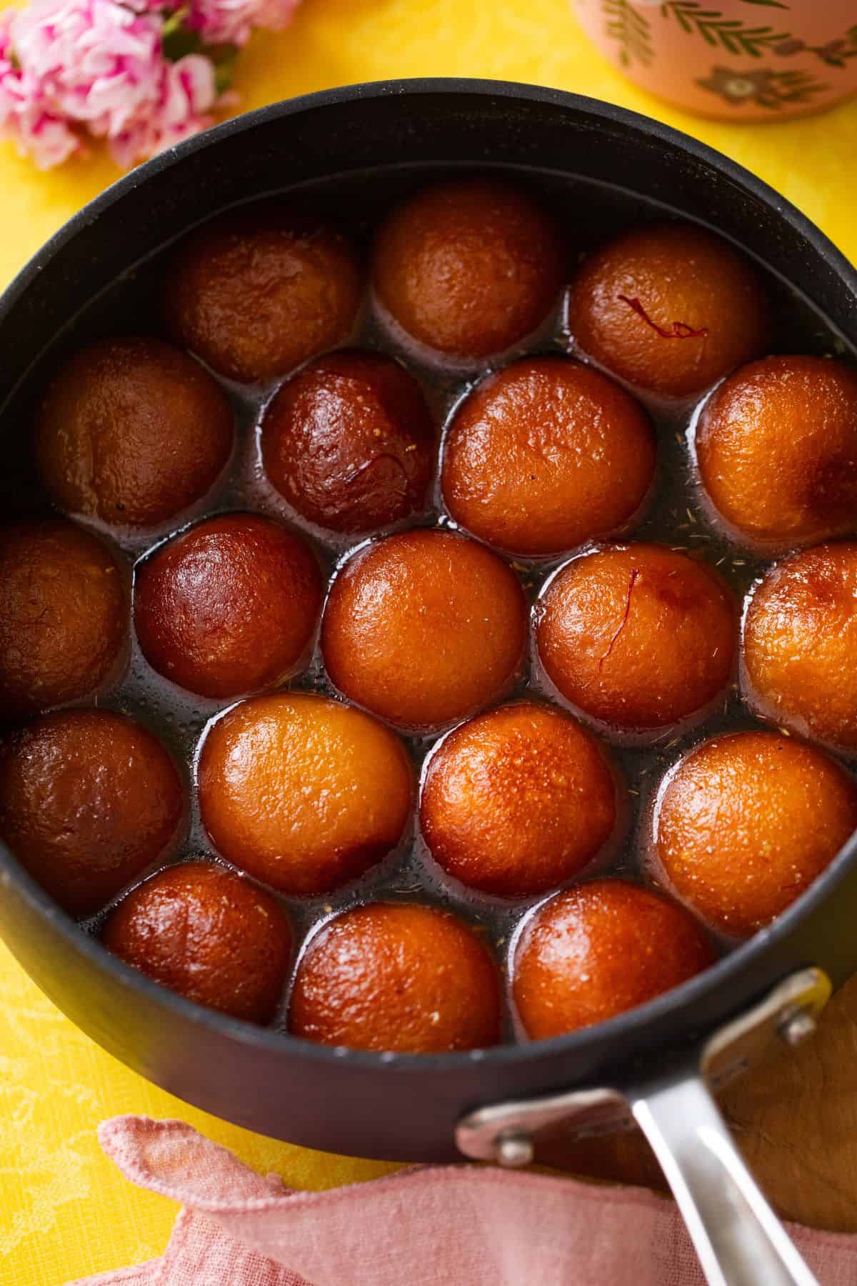 A black pan filled with round, golden-brown gulab jamun soaked in sugar syrup, placed on a yellow surface with a pink cloth and flowers nearby.