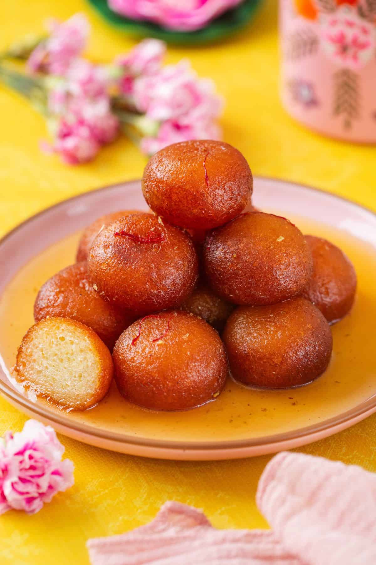 IMG_033711 A plate of golden-brown gulab jamun soaked in syrup, stacked in a pyramid shape, with one piece cut open to show its soft inside. Pink flowers and a floral cup are in the background on a yellow surface.