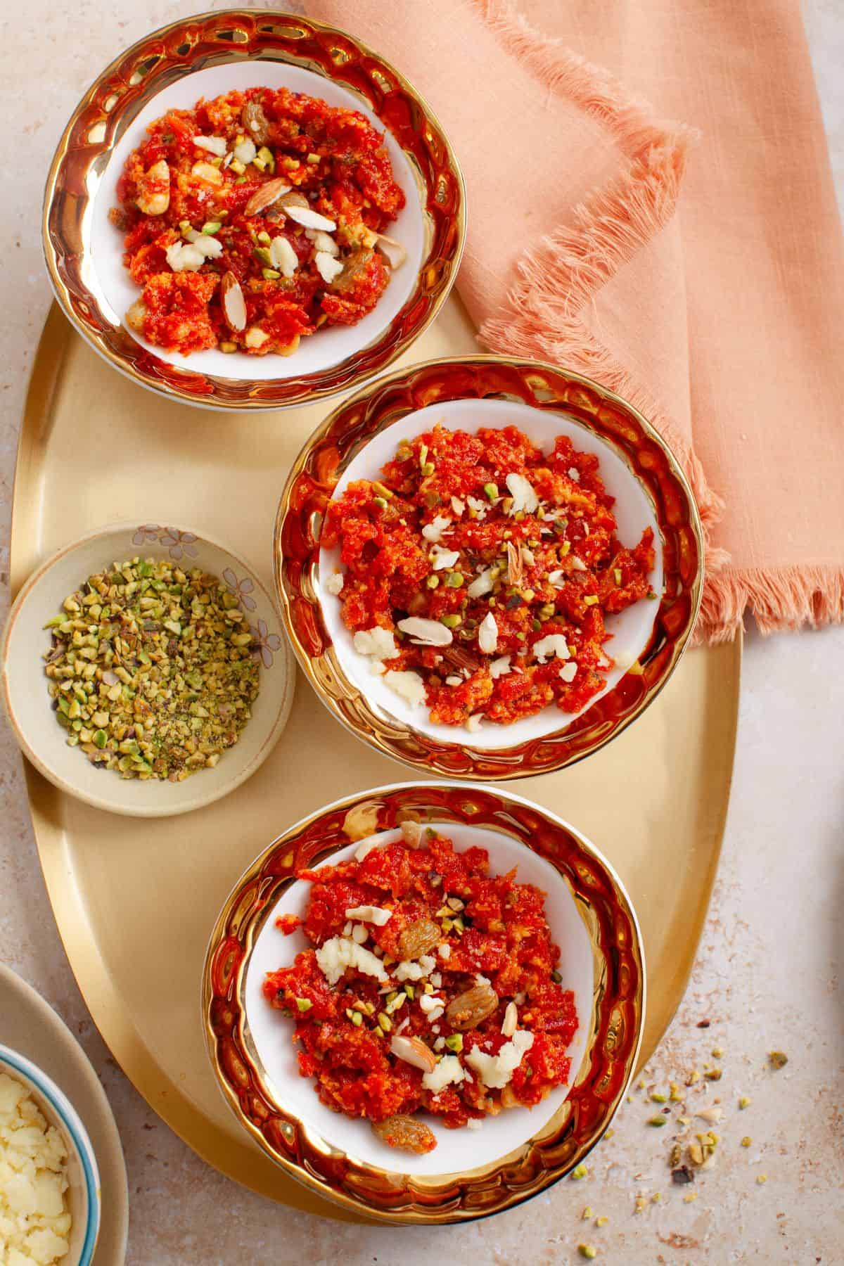Three bowls of gajar halwa placed on a gold platter with a small plate of pistachios.