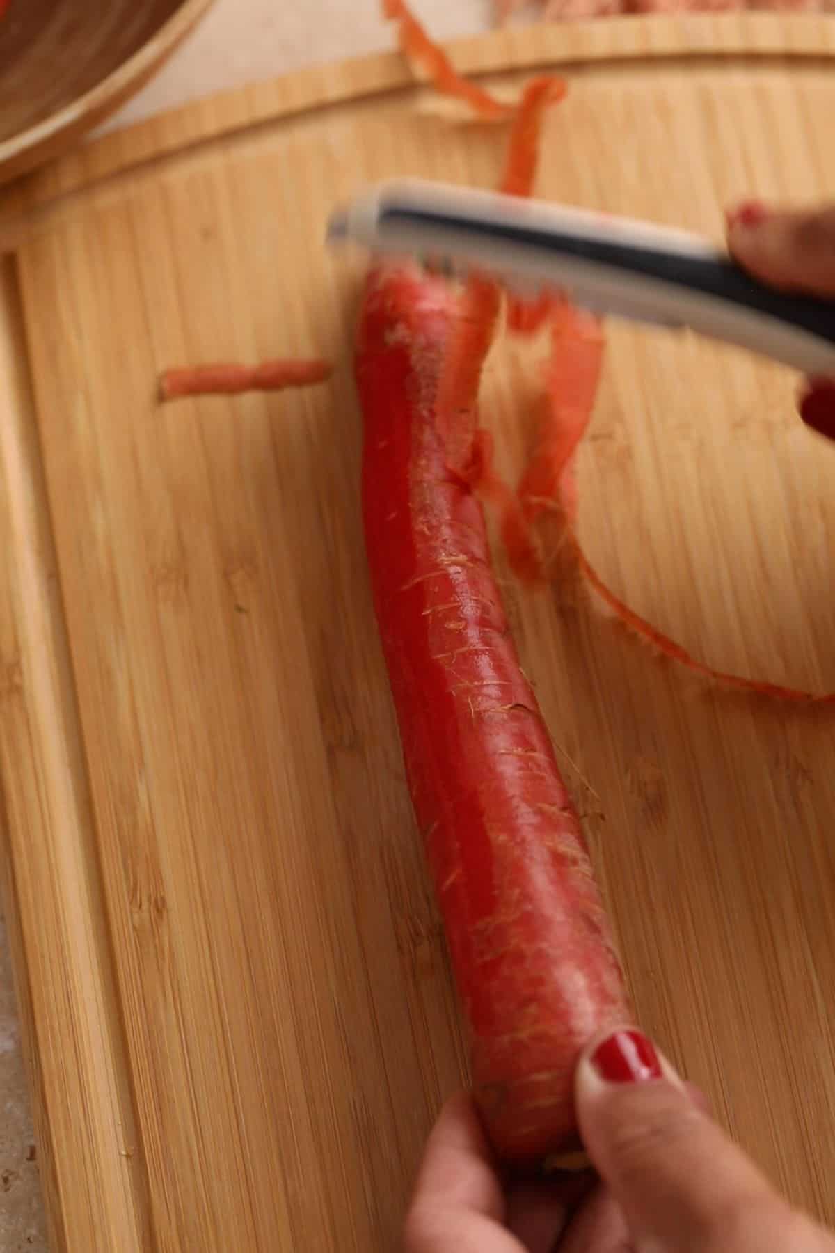 Red carrots are being peeled using a white peeler.