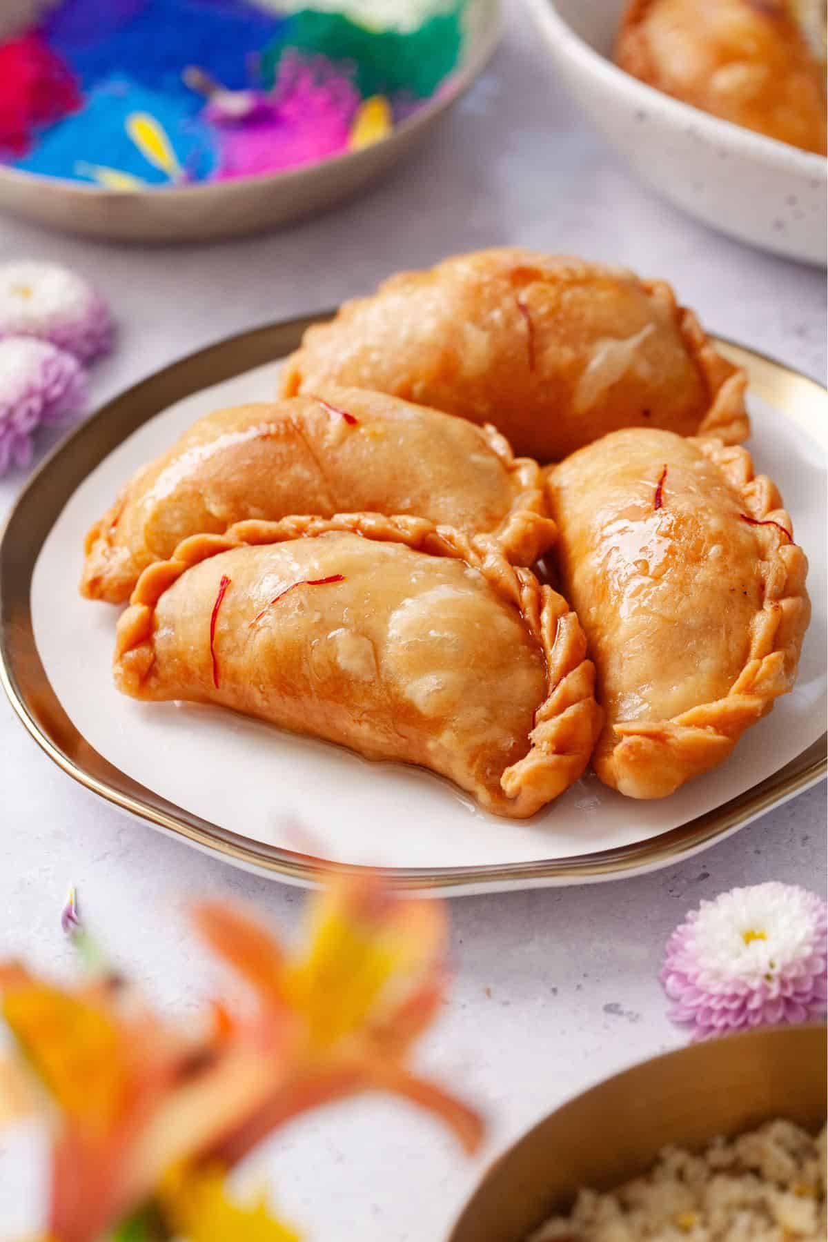 Side image of gujita in a gold and white plate with holi colors in background.