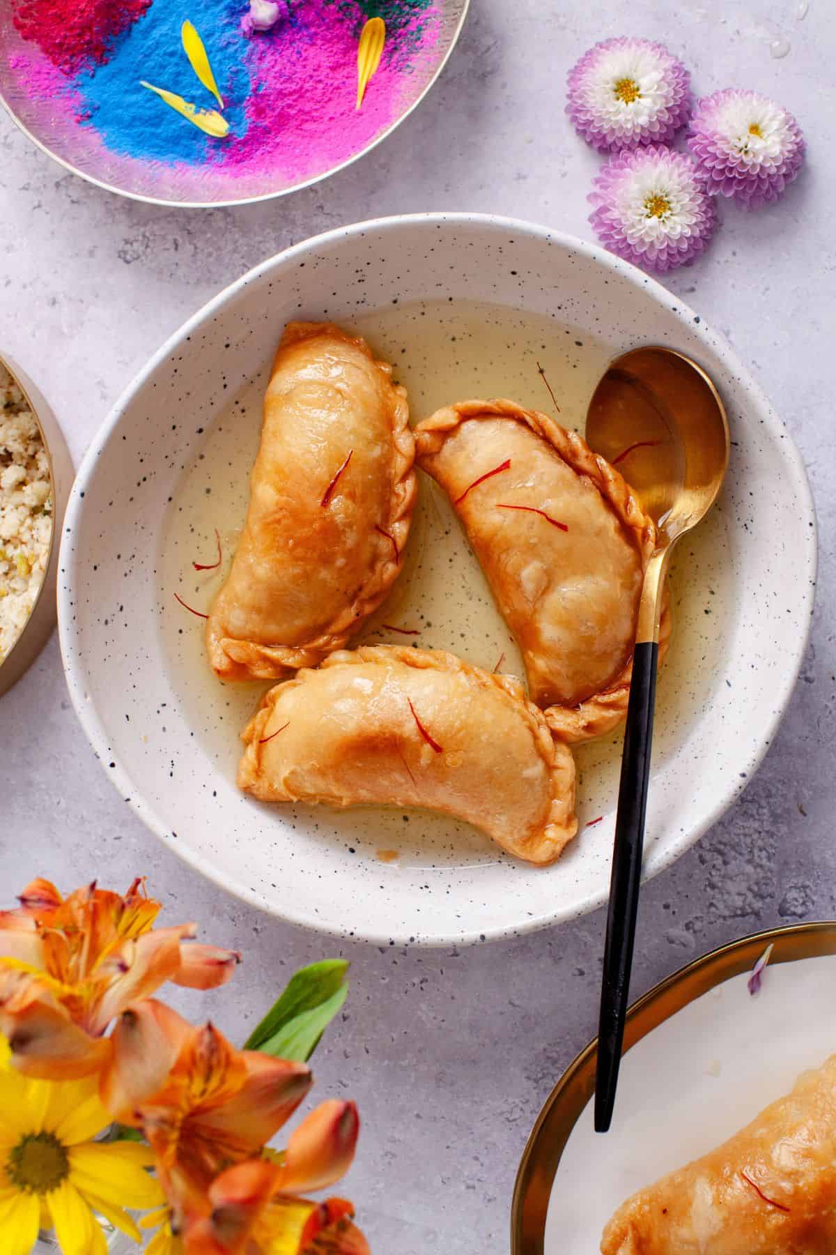 Fried Gujiya soaking in sugar syrup in a ceramic white bowl.