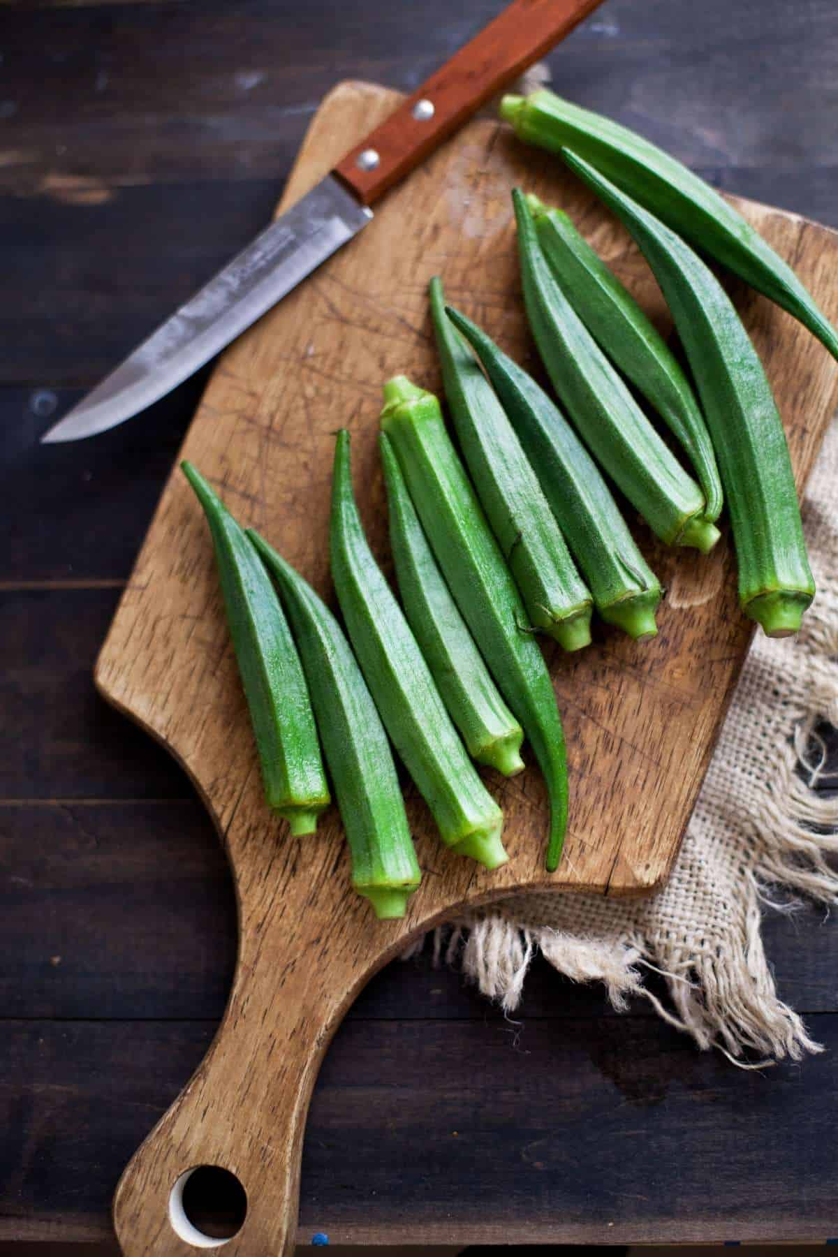 Fresh tender green okra pods on a wooden board with knife.
