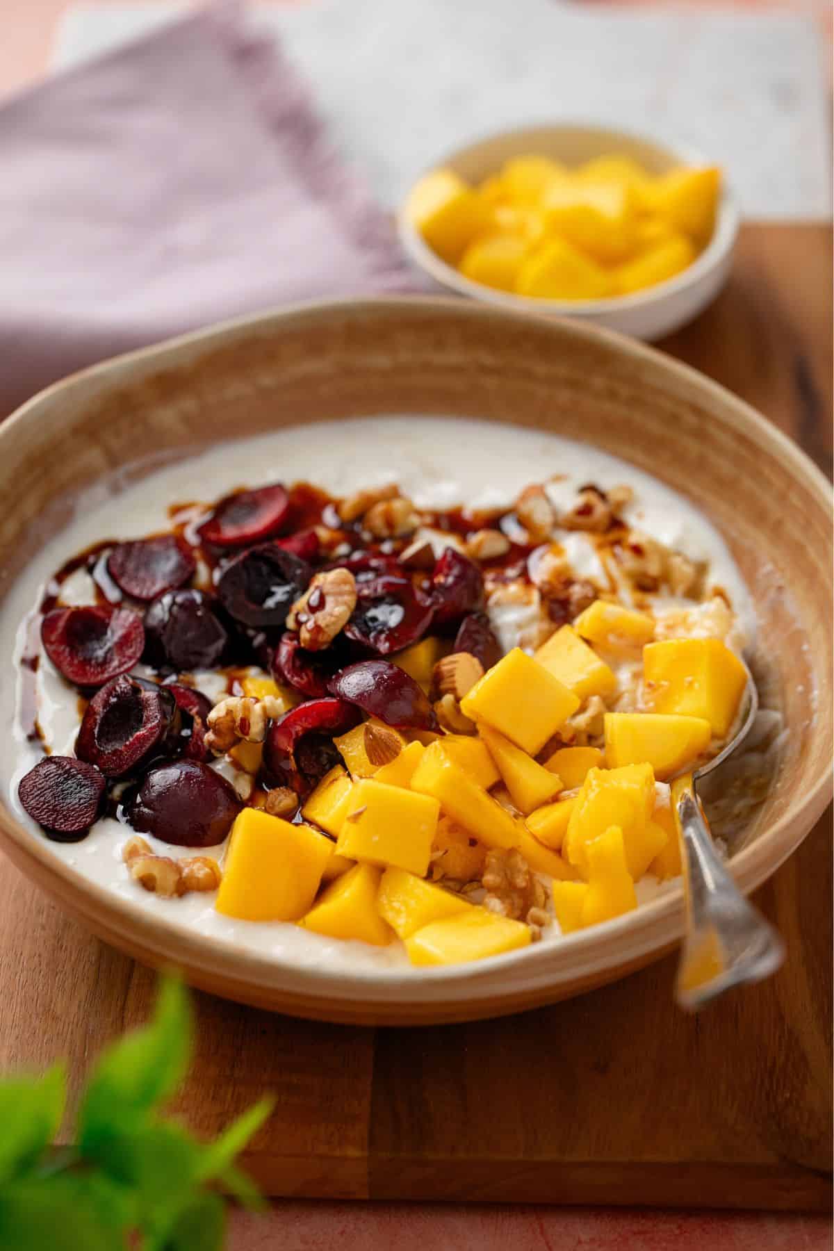 Image of sweet dahi poha served in a beige stone bowl with fresh mango and cherries.