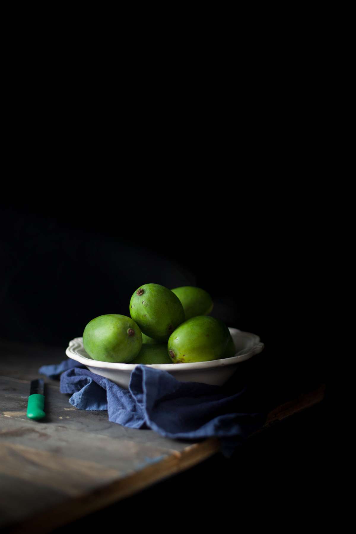 A white bowl with green unripe mangoes against a black background.