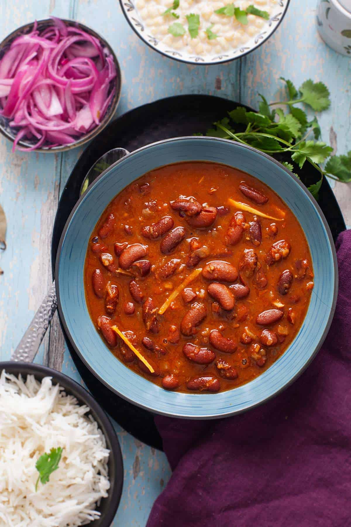 Rajma Recipe servd in a ablue bowl with chawal, raita and pickled onions.