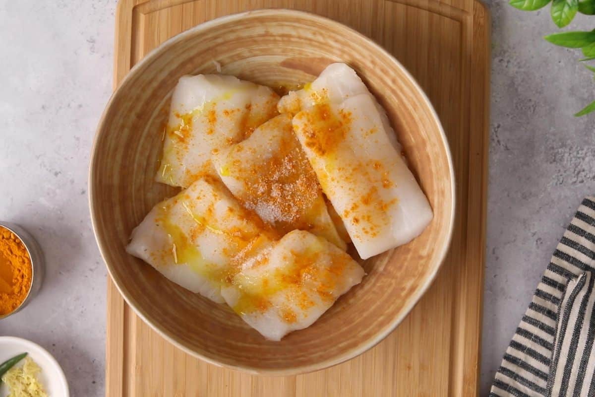 Raw cod pieces placed in a beige bowl are sprinkled with spices.