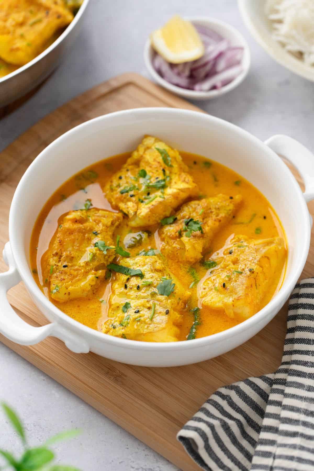 Cod curry in mustard sauce served in a white dish with sliced onions, lemon and rice in background.