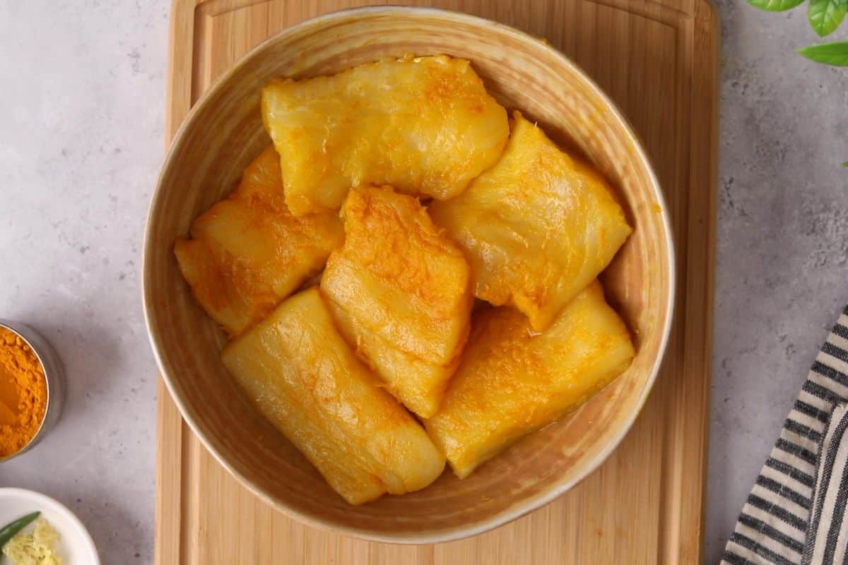 Raw cod pieces placed in a beige bowl are coated in salt, turmeric and mustard oil.
