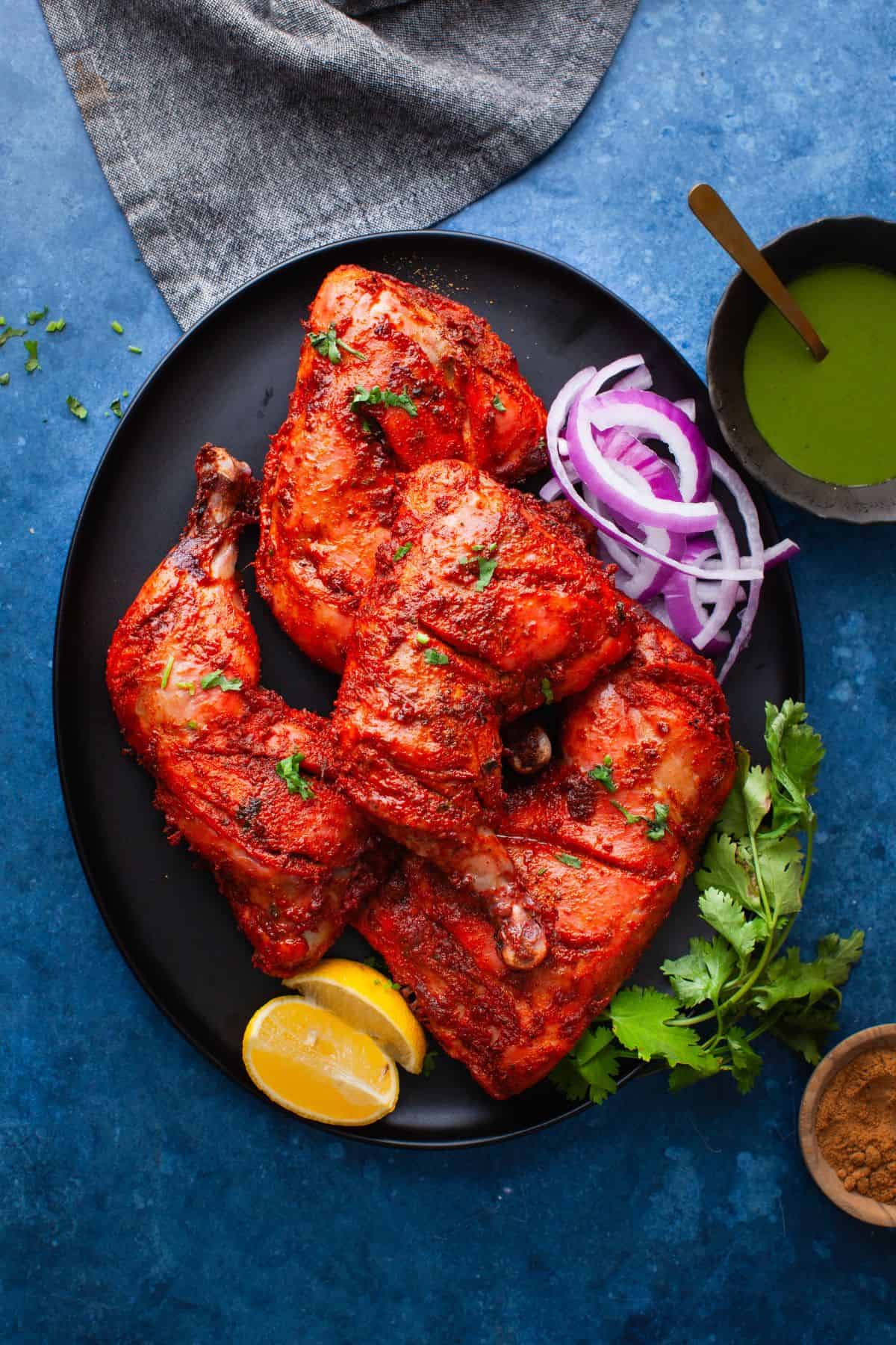 Tandoori chicken quaters place on a black platter with thin sliced onions, lemon and green chutney.