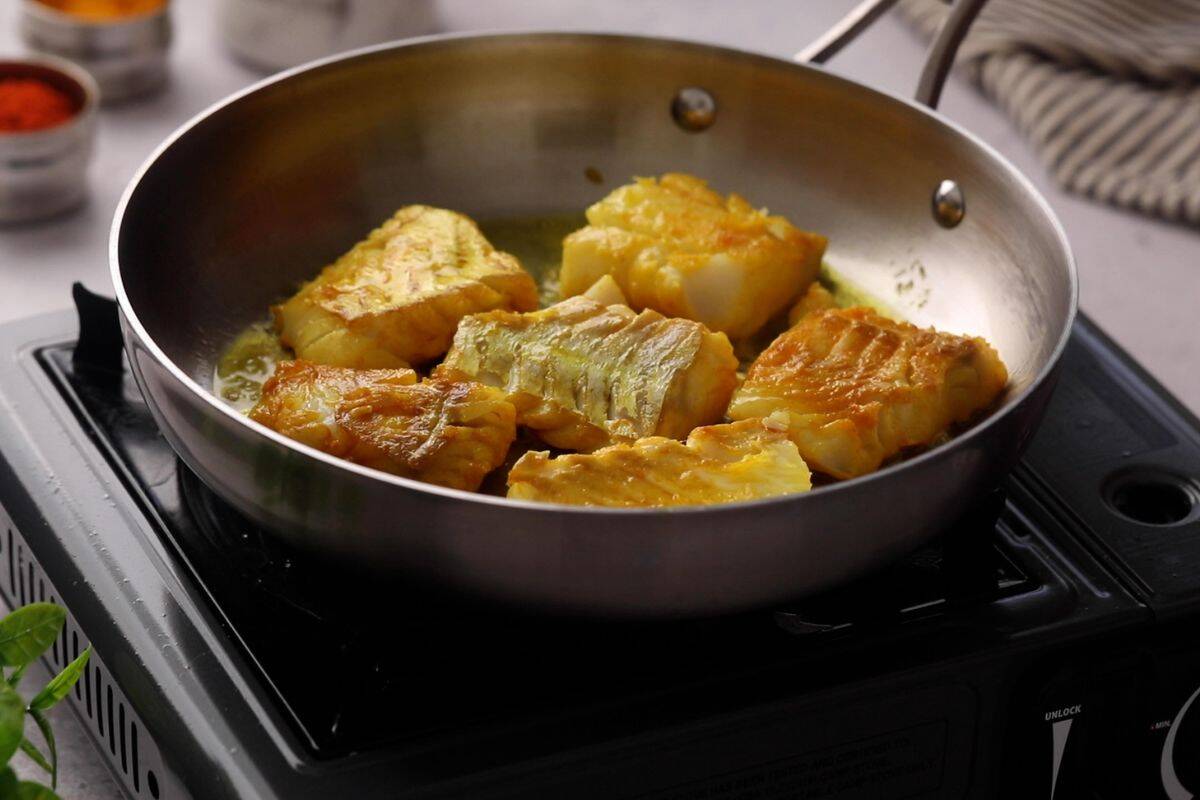 Cod is seared in hot oil in a stainless steel pan.