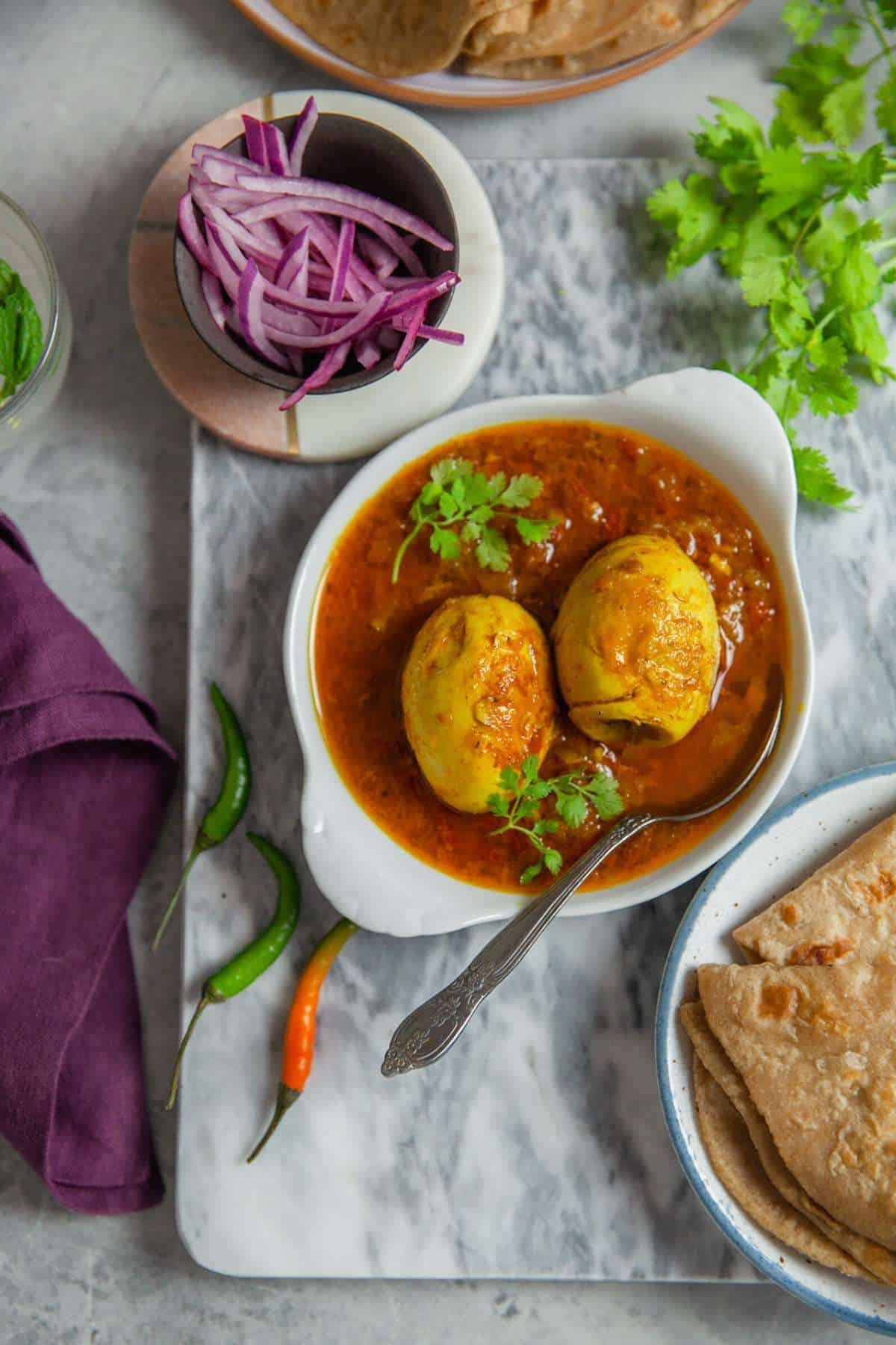 Punjabi egg curry served in a small white dish with pickled onions, indian flatbread and fresh herbs.