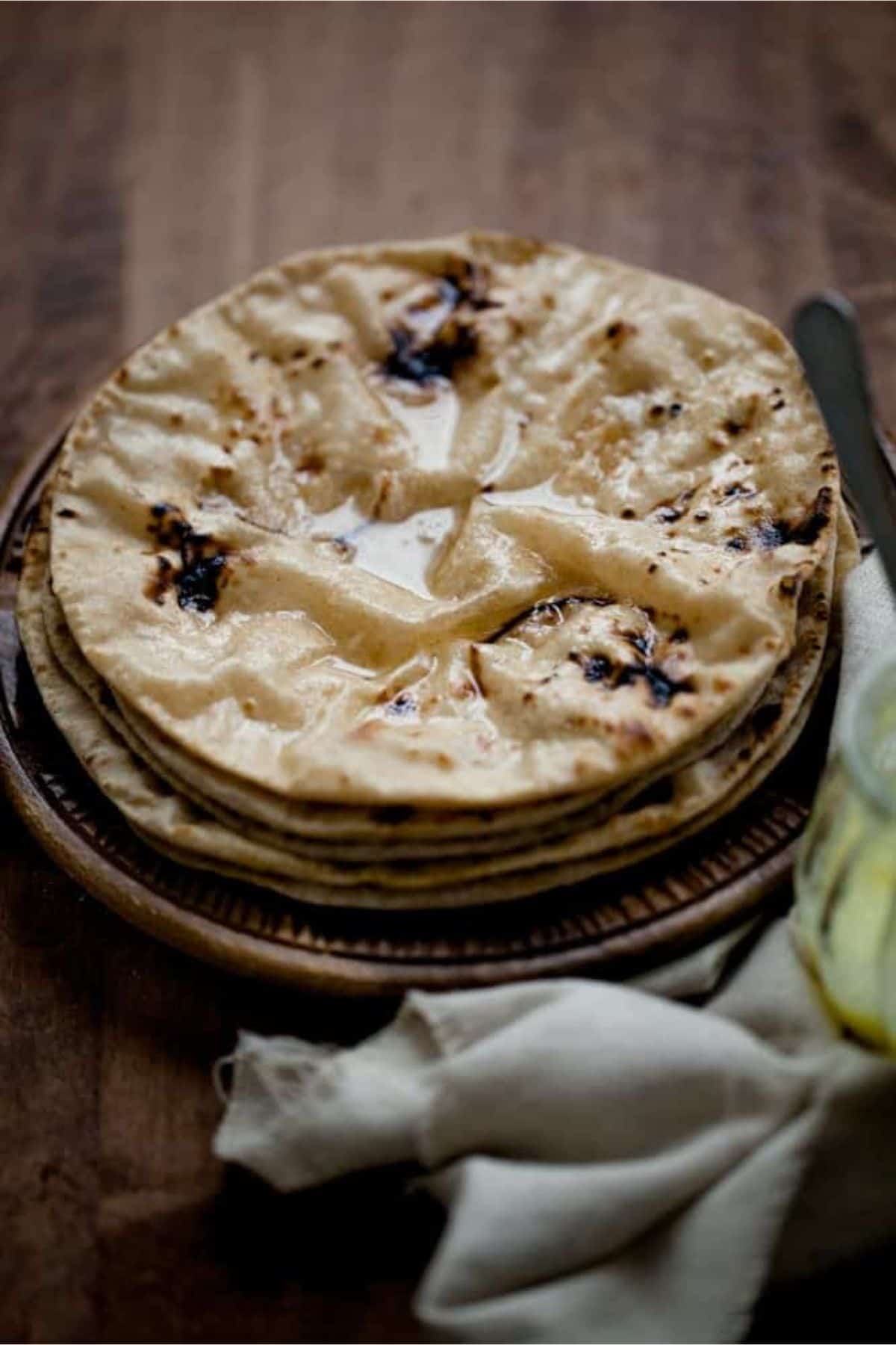 A stack of freshly made chapatis topped with melted ghee sits on a brown plate, with a jar of ghee and a beige cloth napkin beside it on a wooden surface.