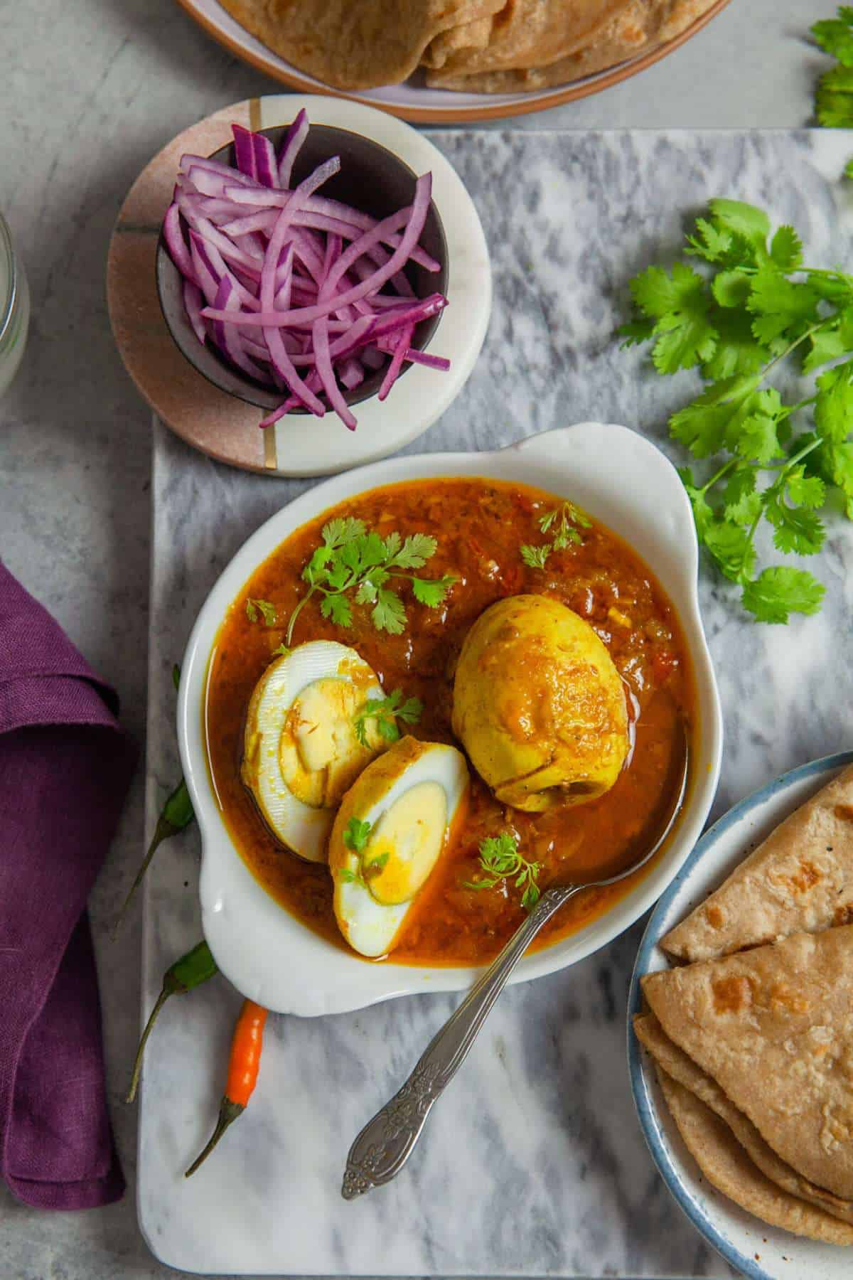 North indian style egg curry showing the halved eggs served in a small white dish with pickled onions, indian flatbread and fresh herbs.