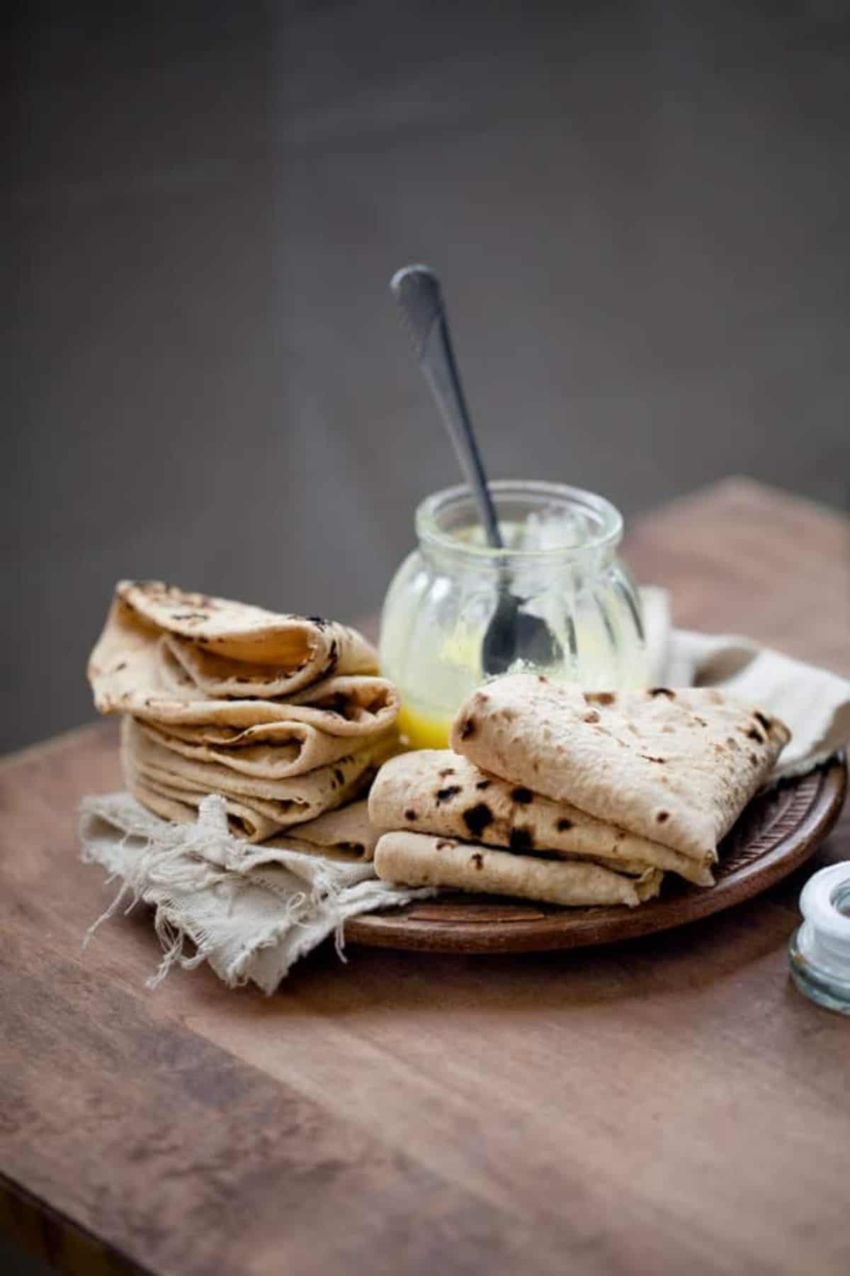 A wooden plate with folded and stacked pieces of roti flatbread sits on a rustic cloth next to a glass jar of yellow clarified butter with a spoon.