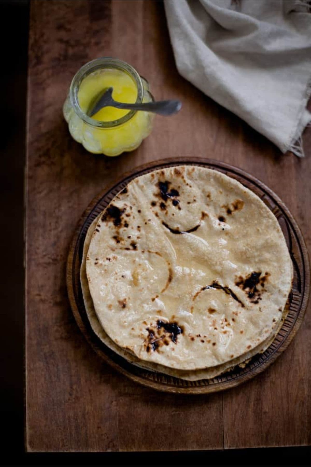 Top view of stack of roti on a wooden plate with a small jar of ghee.