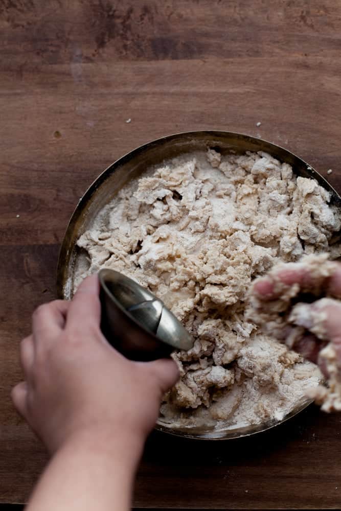 Mixing flour in a metal plate on a wooden surface, one hand holding a metal cup and the other hand kneading the crumbly flour mixture.