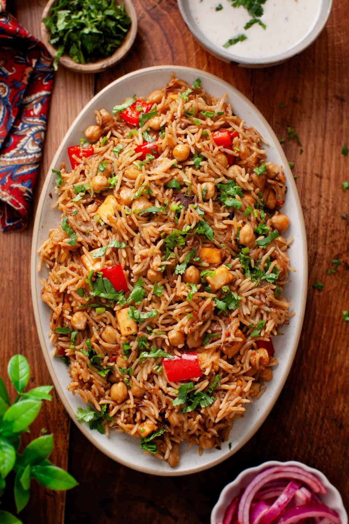An oval platter of chana pulao with chickpeas, diced red bell peppers, herbs, and spices sits on a wooden table.