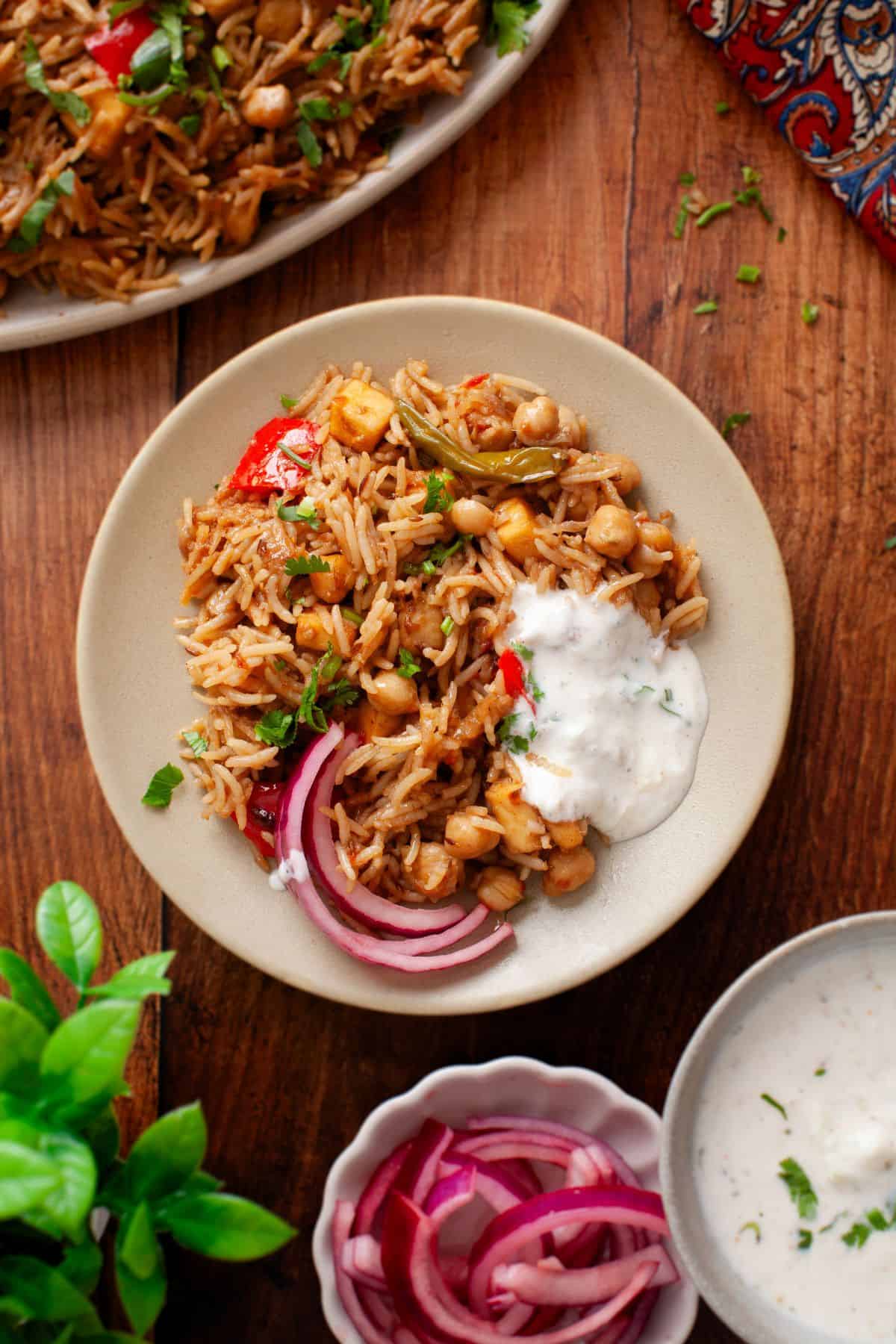 A plate of chickpea pulao garnished with fresh herbs, served with sliced red onions and a dollop of yogurt, sits on a wooden table. 