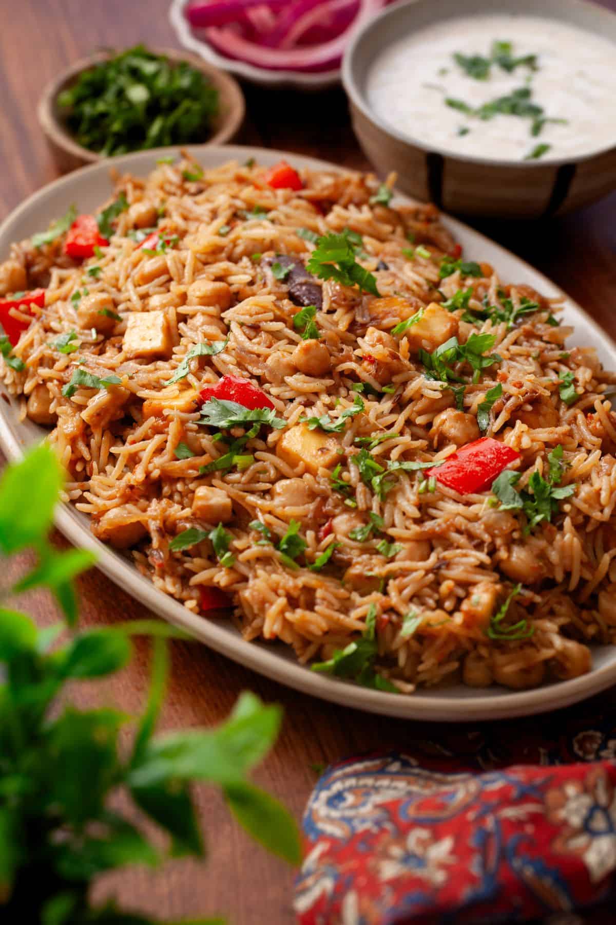 A side image of plate of chana pilaf garnished with fresh cilantro, served on a wooden table with bowls of yogurt sauce and sliced onions in the background.