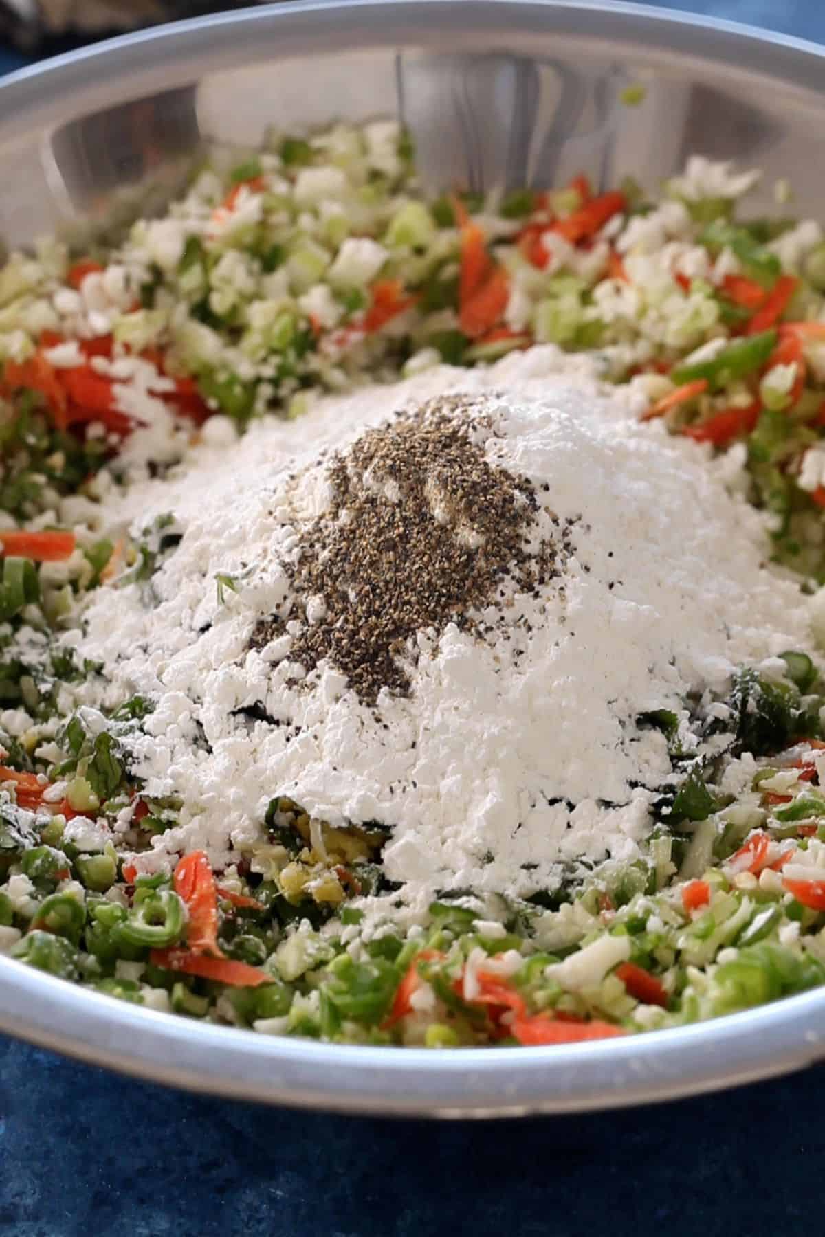 A large metal bowl containing a mixture of chopped vegetables, flour, and black pepper. 