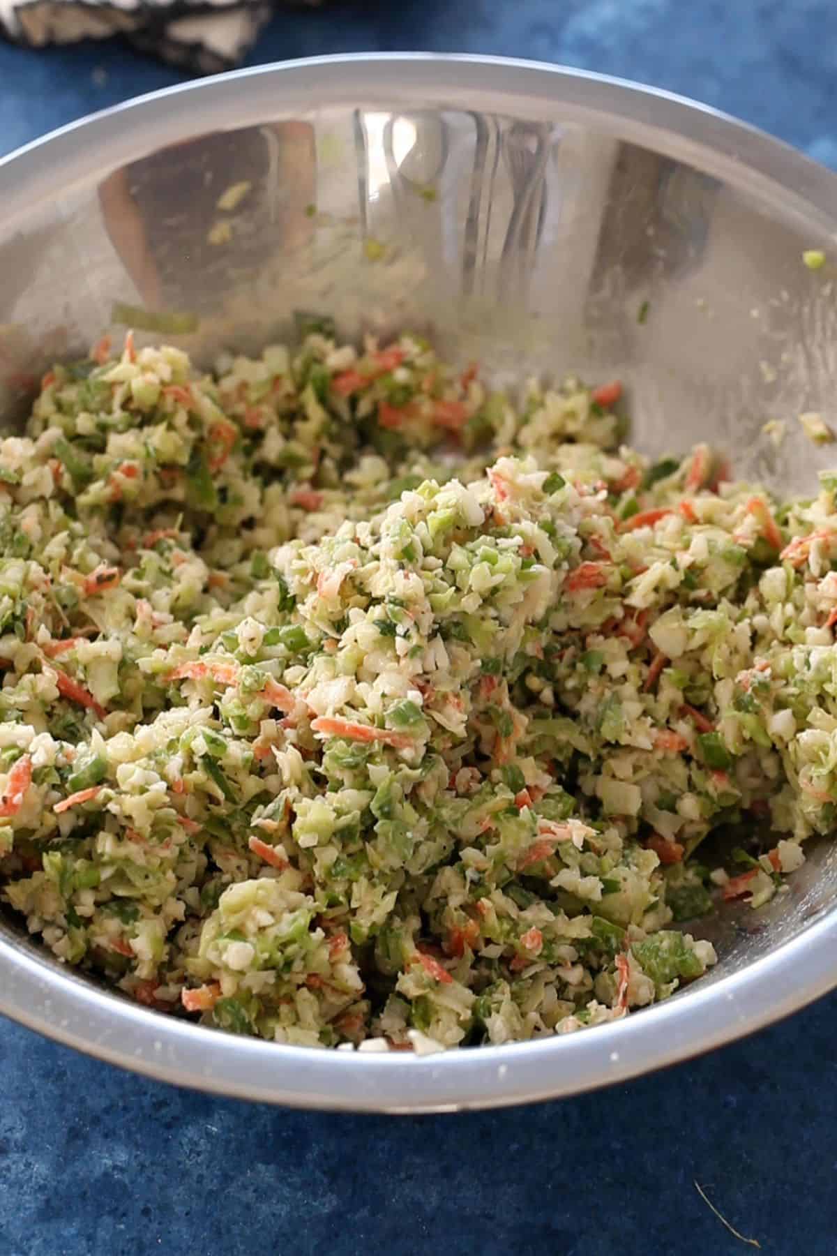 A stainless steel bowl filled with finely chopped vegetables, including carrots, cabbage, and greens, resembling coleslaw, rests on a blue countertop.
