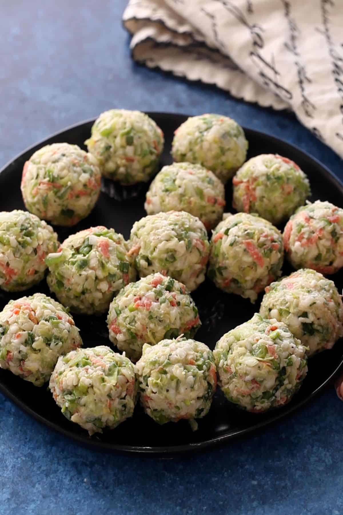A black plate holds neatly arranged, round dumplings made from finely chopped vegetables such as carrots, green onions, and cabbage.