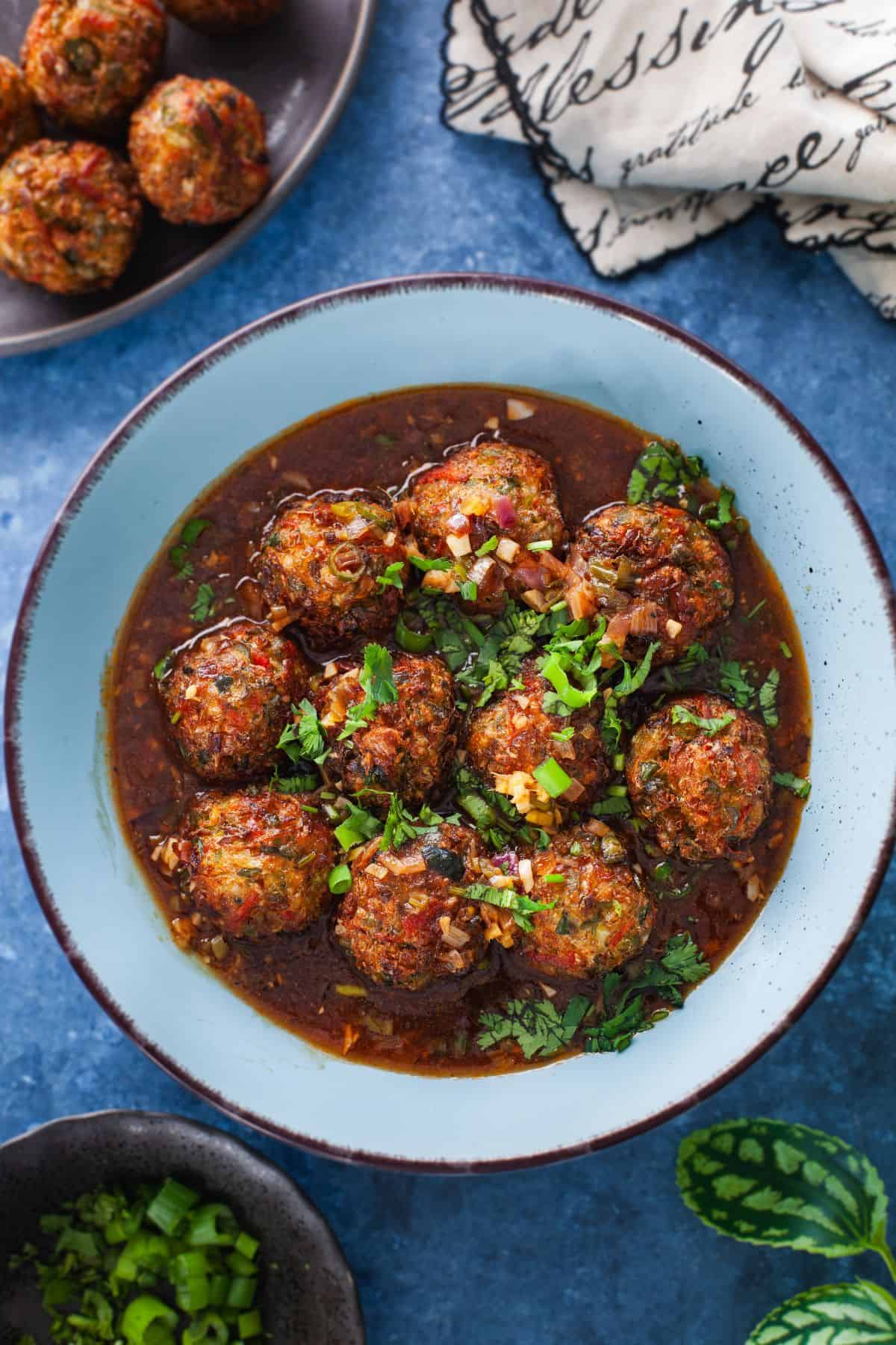 A bowl of spicy vegetable manchurian balls in a rich, dark sauce, garnished with chopped green onions and cilantro.