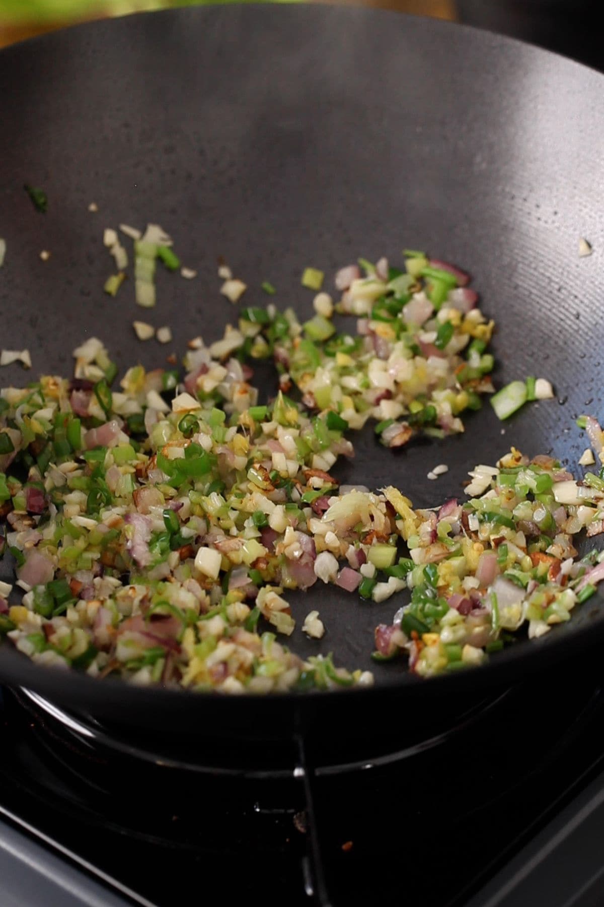 A close-up of a wok on a stovetop, with chopped onions, garlic, green peppers, and herbs being sautéed.
