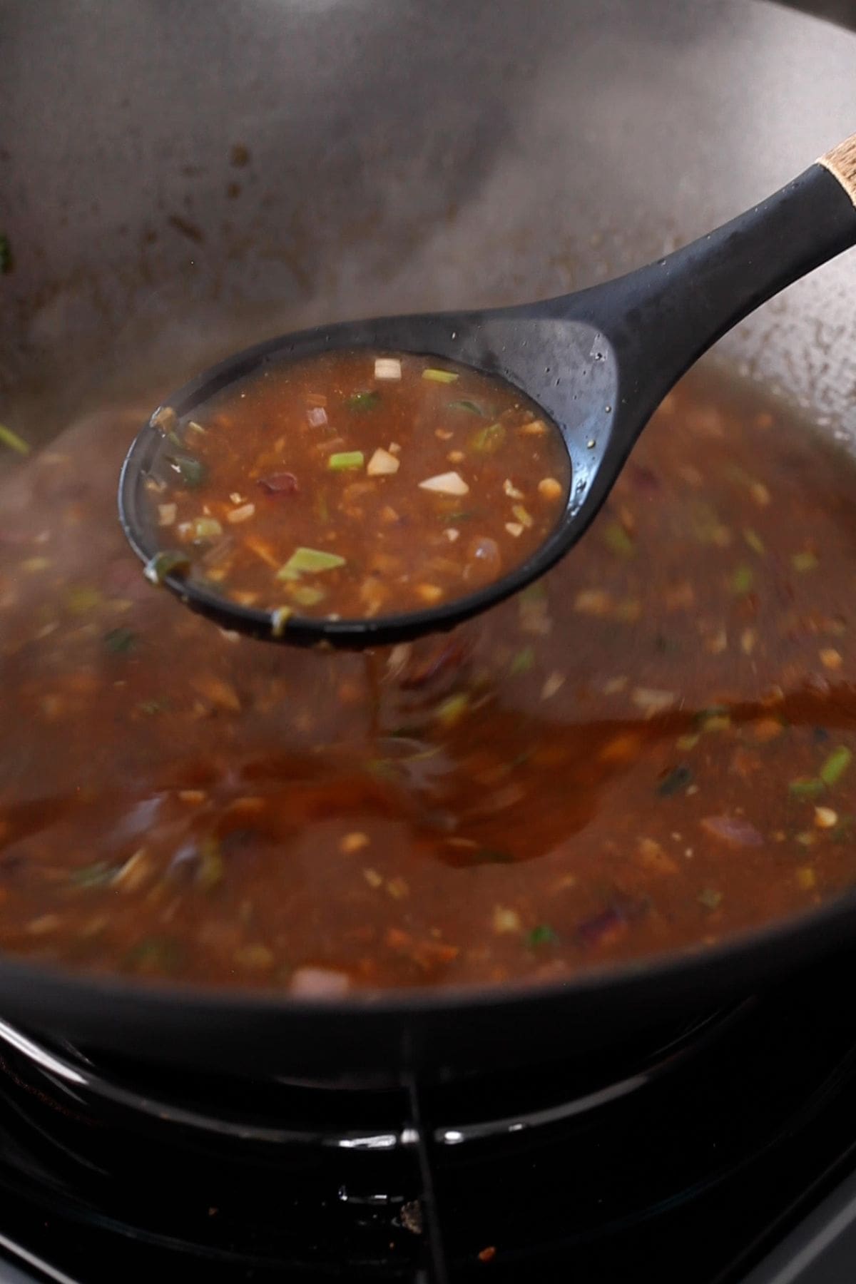 A black ladle scooping a rich, brown manchurian gravy from a steaming wok.