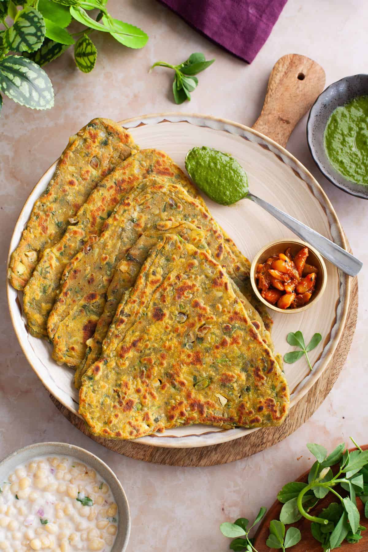 Methi paratha served on a beige plate with pickle, raita and green chutney.