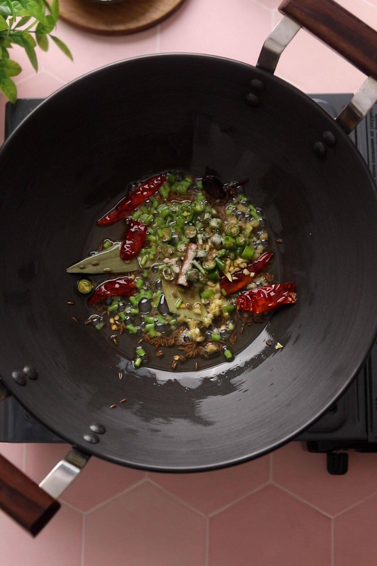 Whole spices and aromatics are frying in ghee in a black kadai.