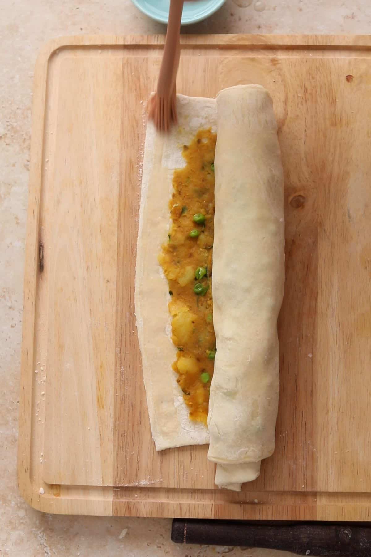 Rolling up pastry spread with a  potato and green pea filling on a wooden cutting board.