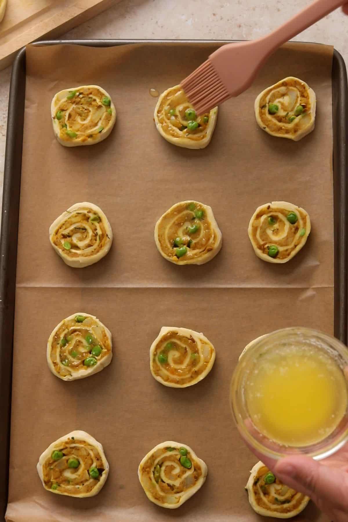 Brushing melted butter to unbaked savory pinwheel rolls with peas and spices, arranged on a parchment-lined baking sheet.