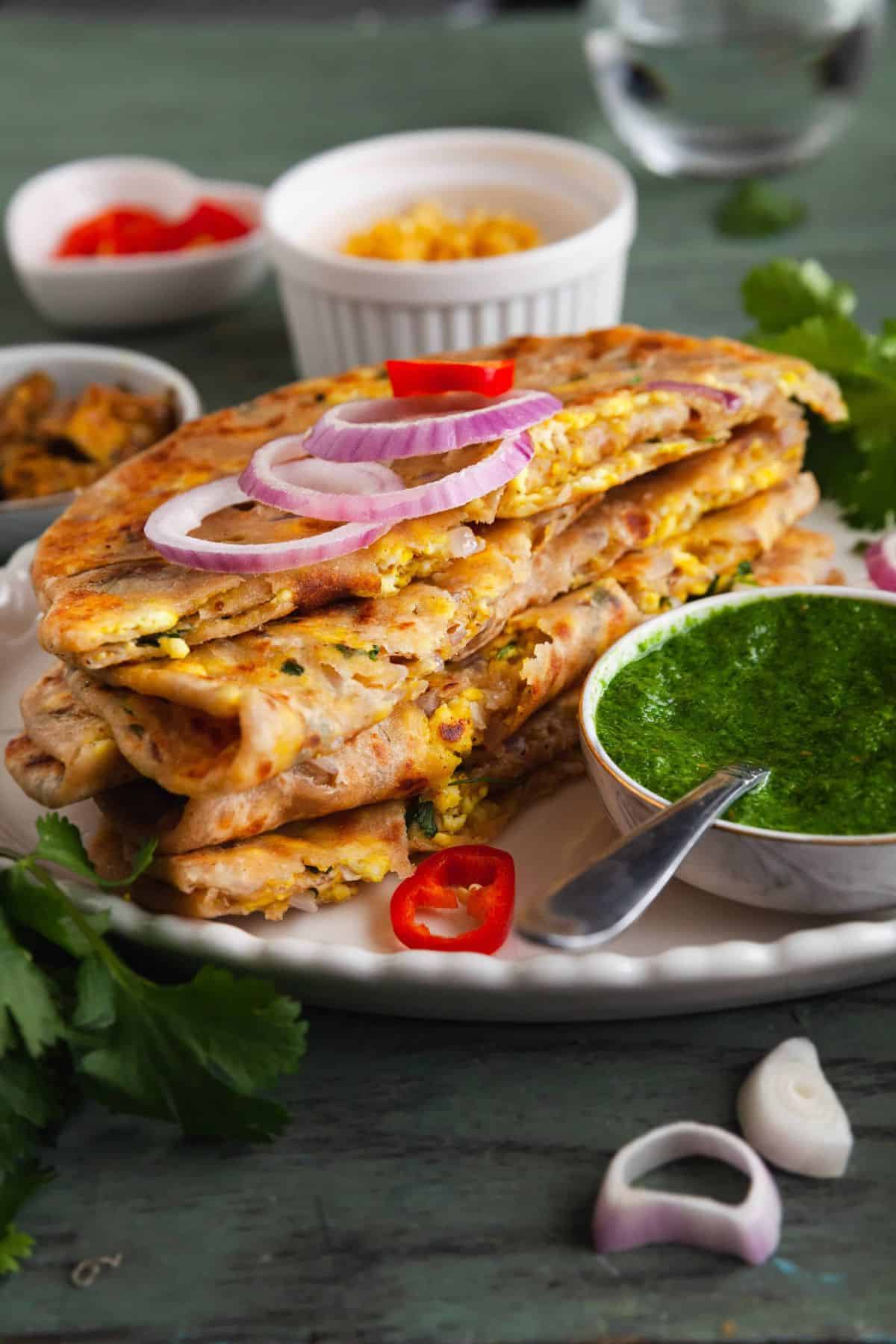 Stack of paneer parathas served with green chutney on a white platter. 