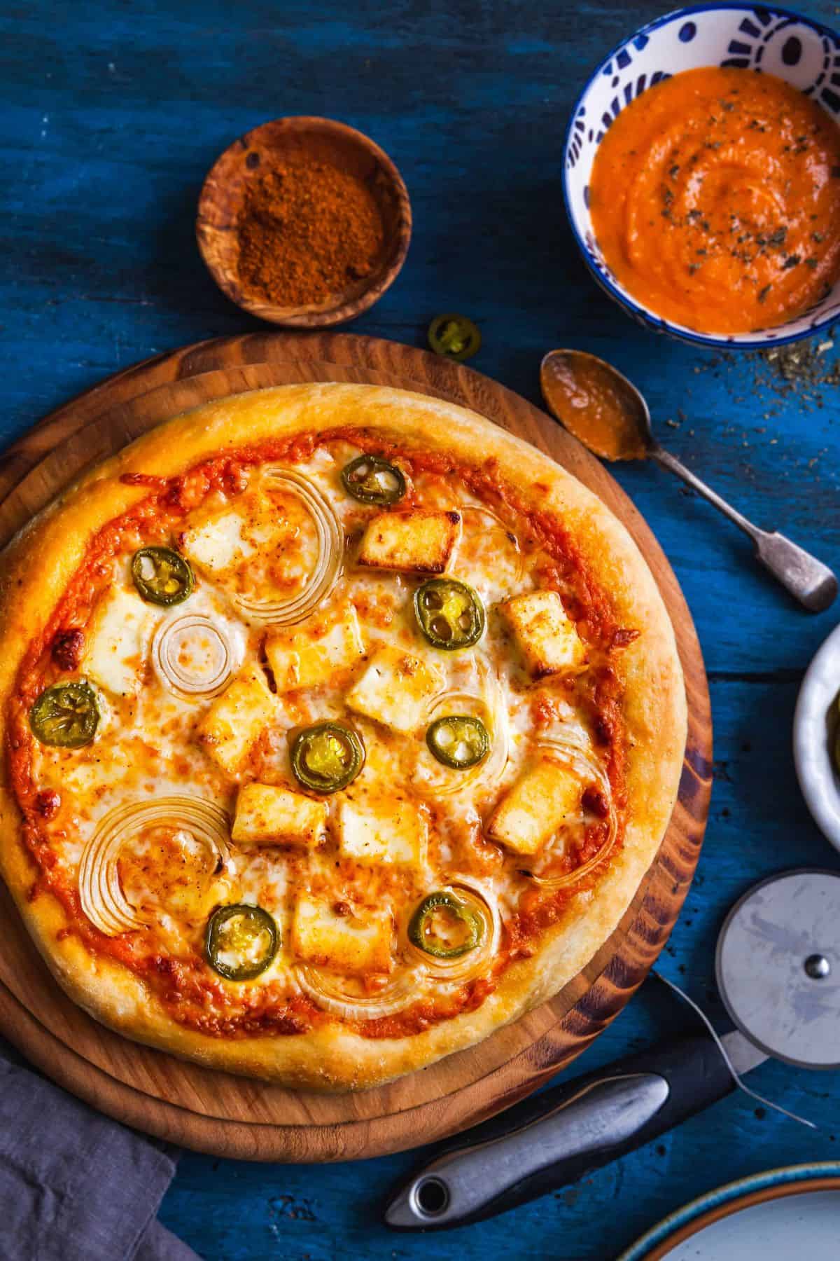Paneer Pizza on a wooden board with a bowl of makhani sauce. 