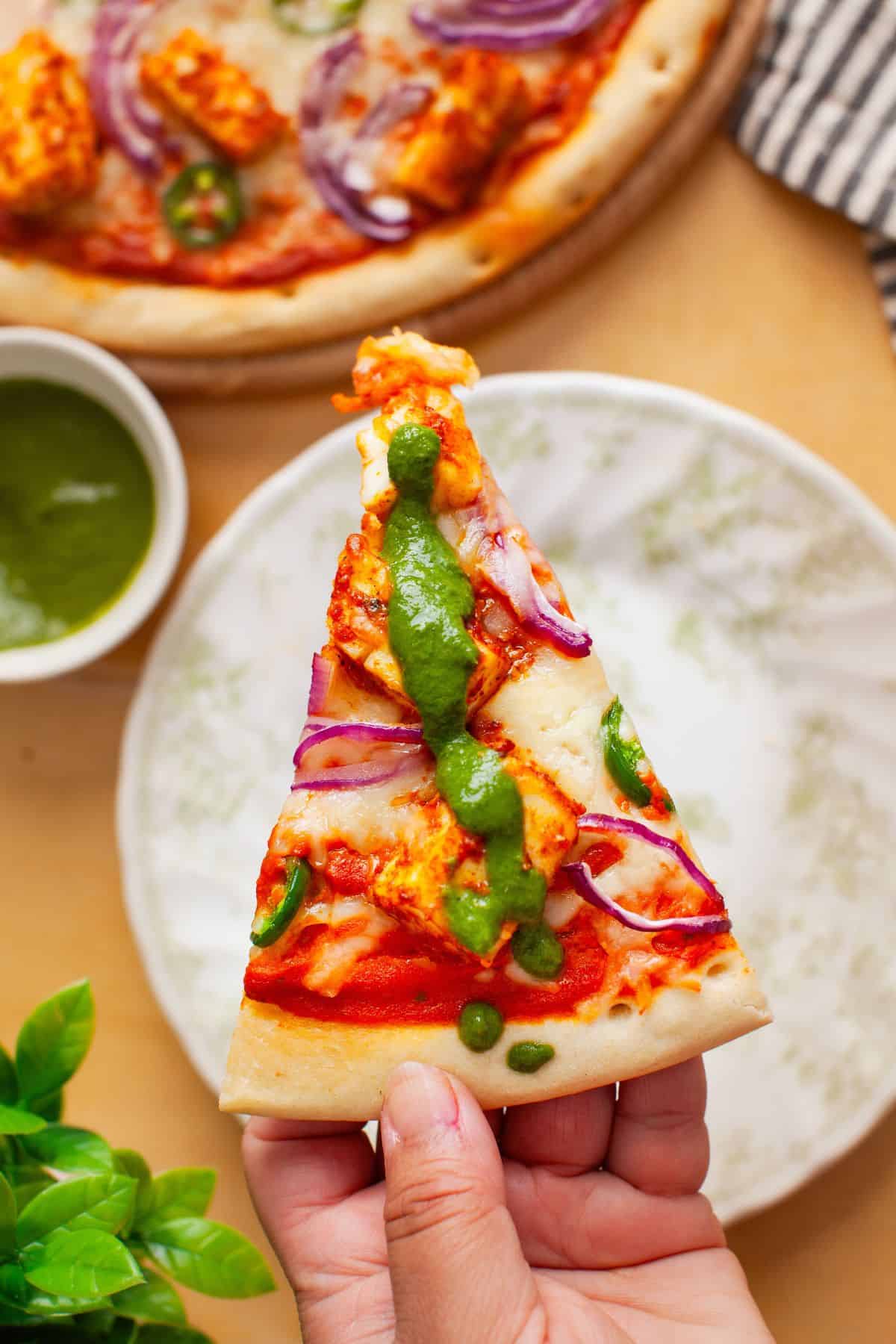 Slice of paneer pizza in hand with drizzle of green chutney.
