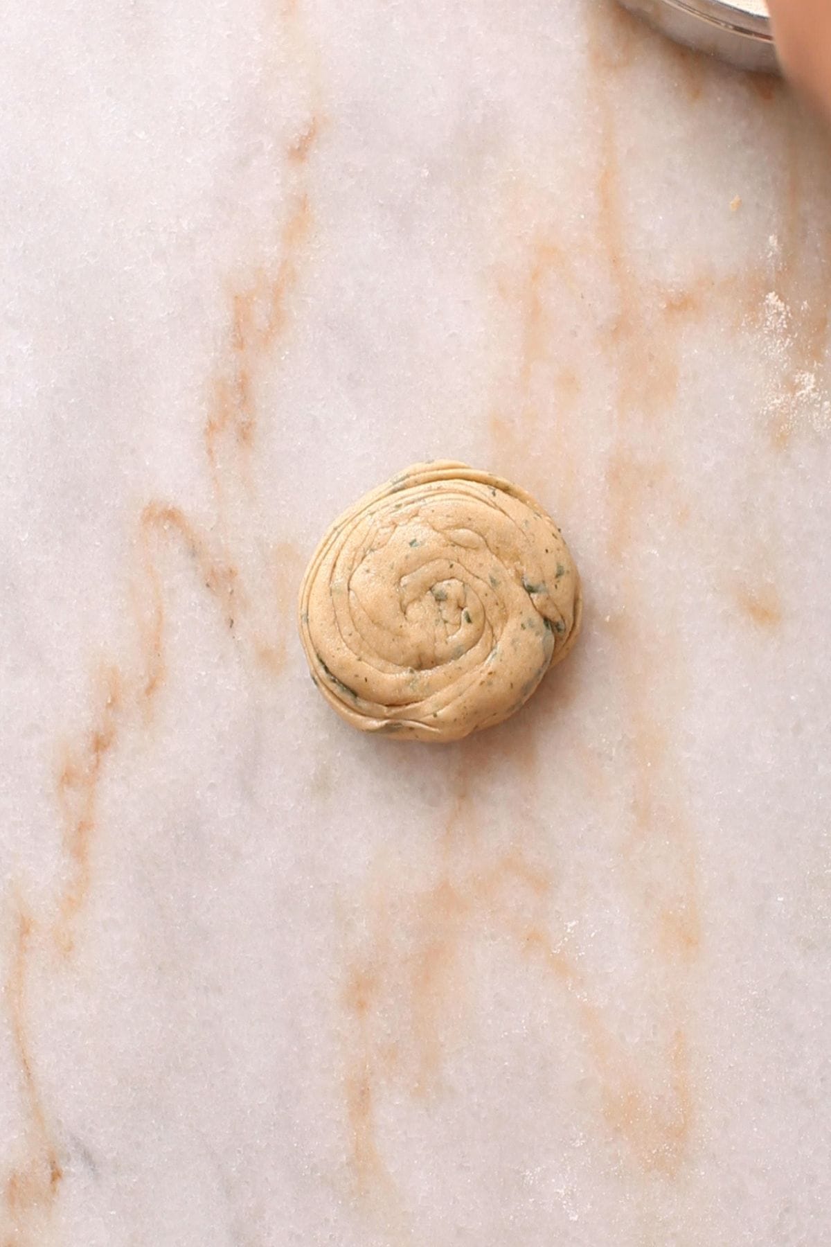 A small, round haped spiral of dough sits on a lightly floured marble surface. The dough has visible layers and specks, suggesting it contains herbs or spices.