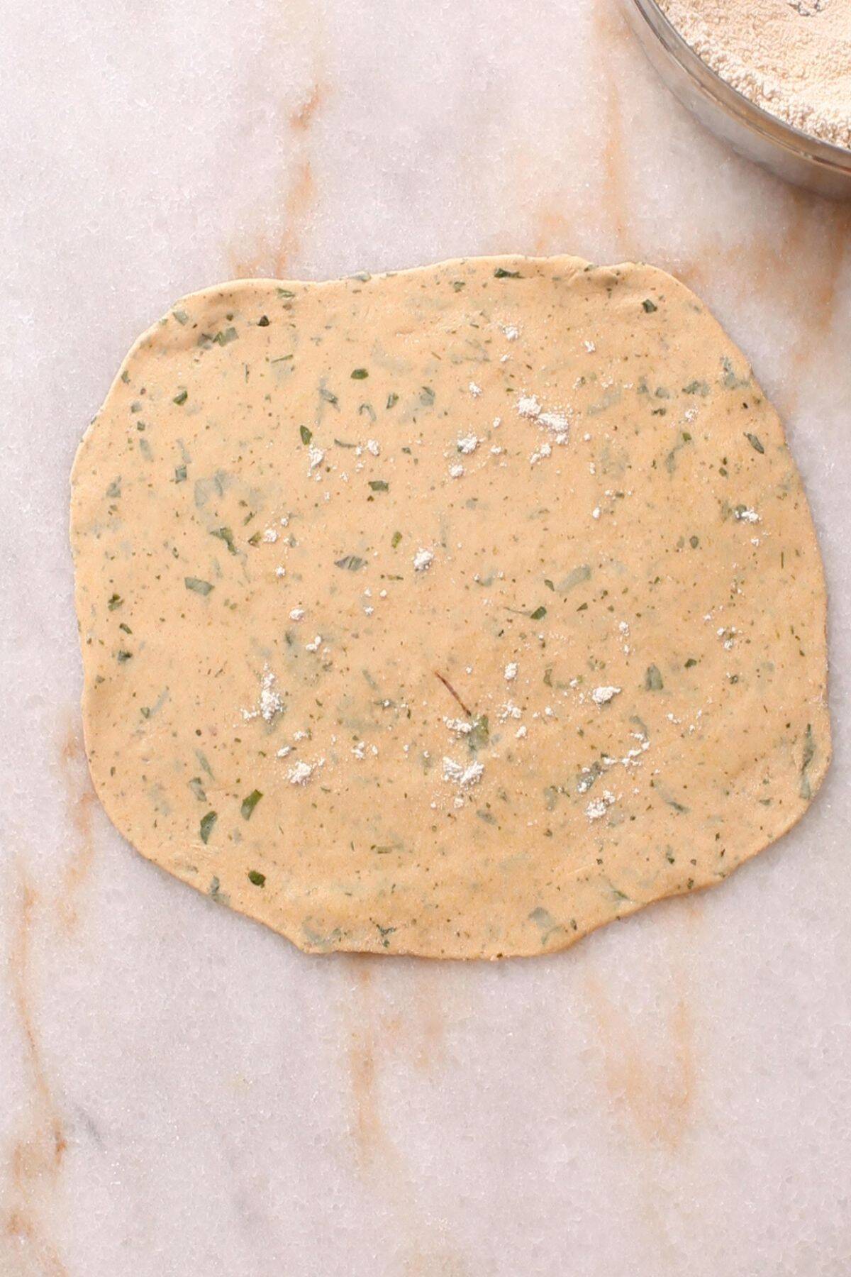 A thinly rolled-out, round piece of dough with herbs and spices is spread on a marble surface, lightly dusted with flour. A bowl with more flour is partially visible in the upper right corner.