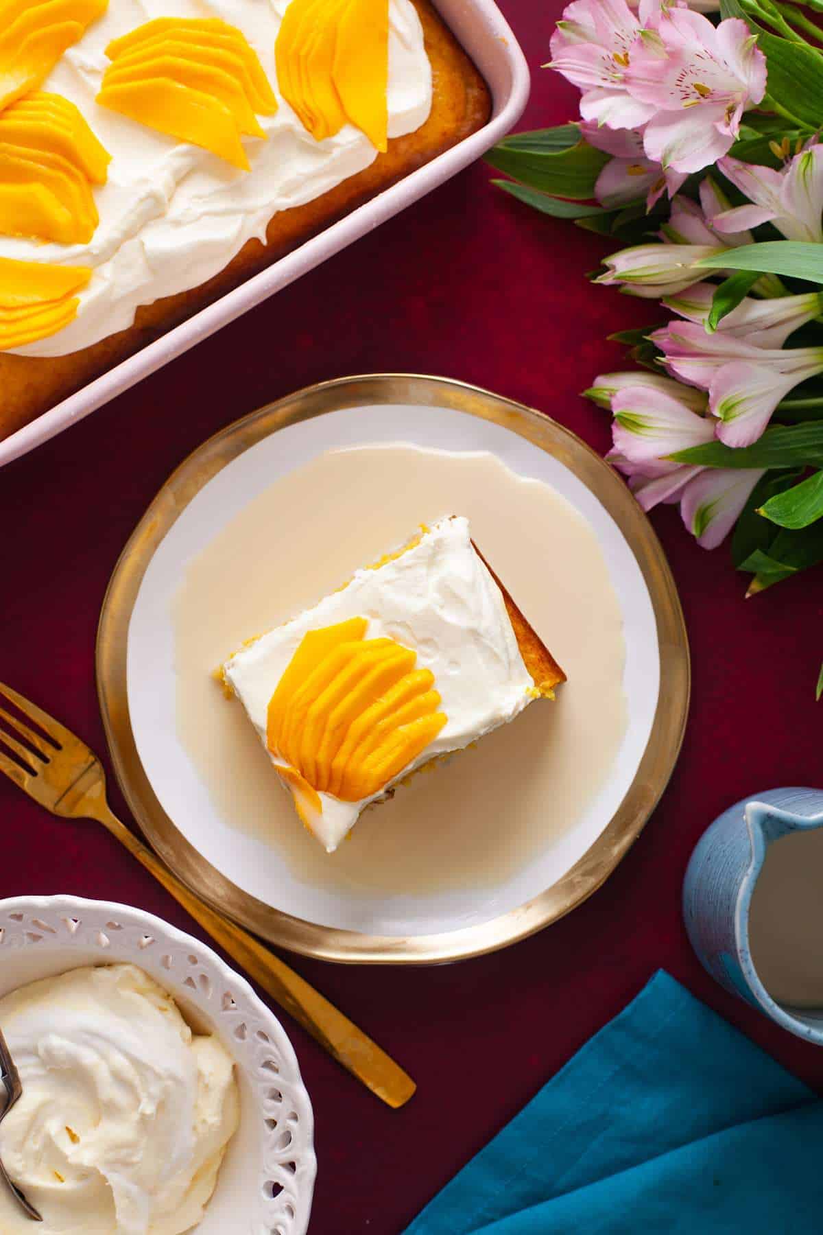 Top view of a slice of tres leches cake topped with fresh sliced mangoes, whipped cream and served with a a3 milk mixture in a gold rimmed plate.