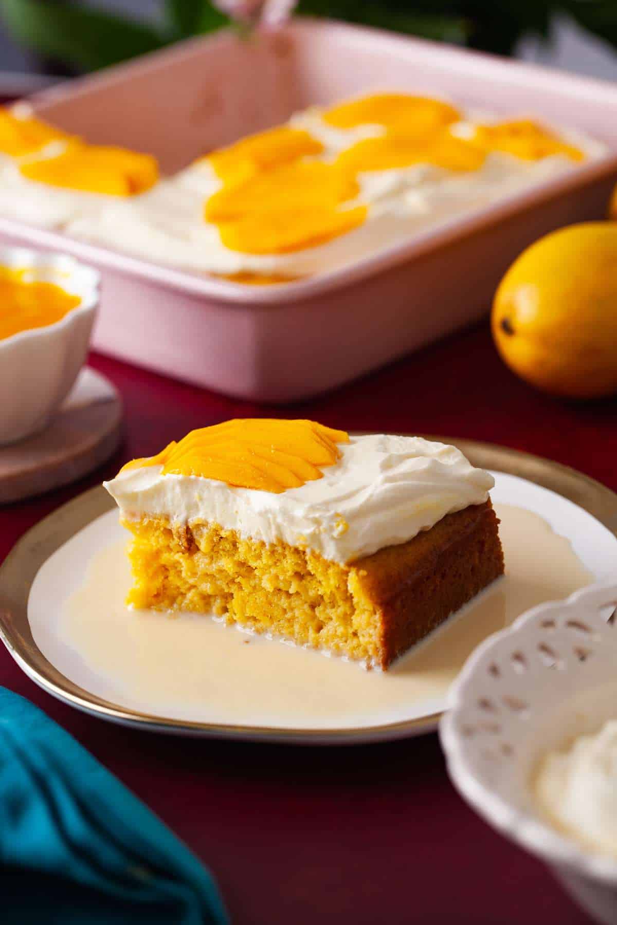 Side view of a sliced of mango tres leches cake with mango puree, whipped creamed fresh mangoes in the frame. 