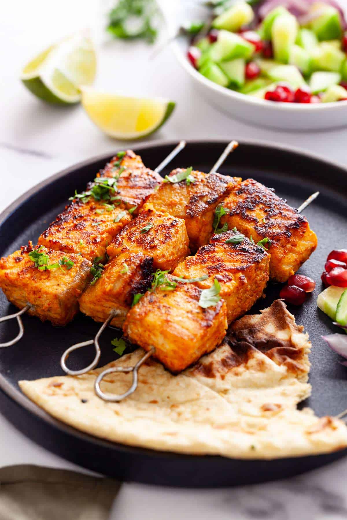 Tandoori Salmon Tikka Skewers on a black plate with roti and cucumber salad.
