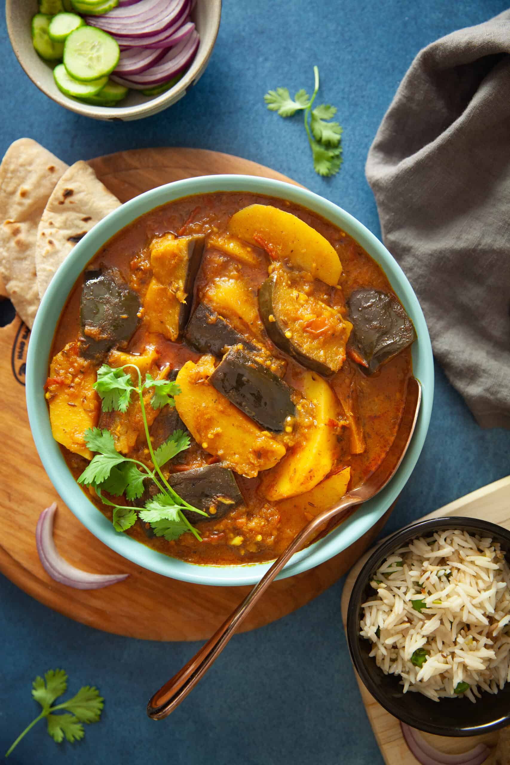 Baingan Aloo in a a blue bowl served with flatbread and rice.